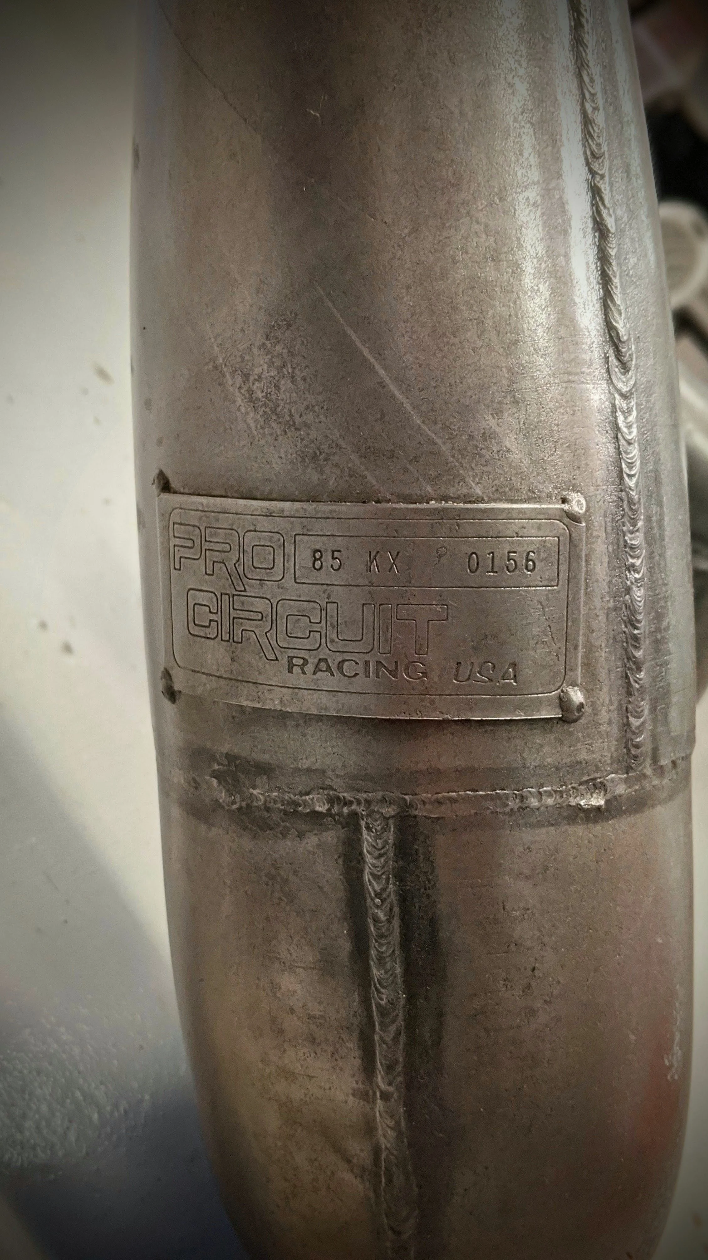 Close-up of a metal racing bicycle component with a metal plate that reads PRO CIRCUIT RACING USA, and includes the numbers 85KX 0156.