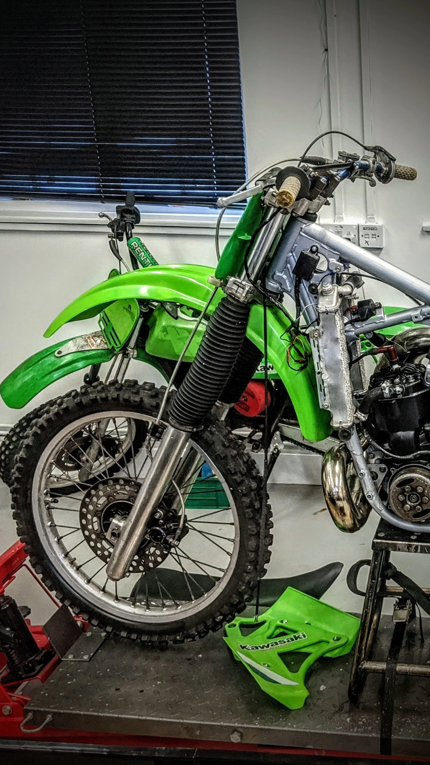 A partially disassembled green Kawasaki dirt bike on a workbench, with the front tire and fender visible, and the front suspension and engine exposed.