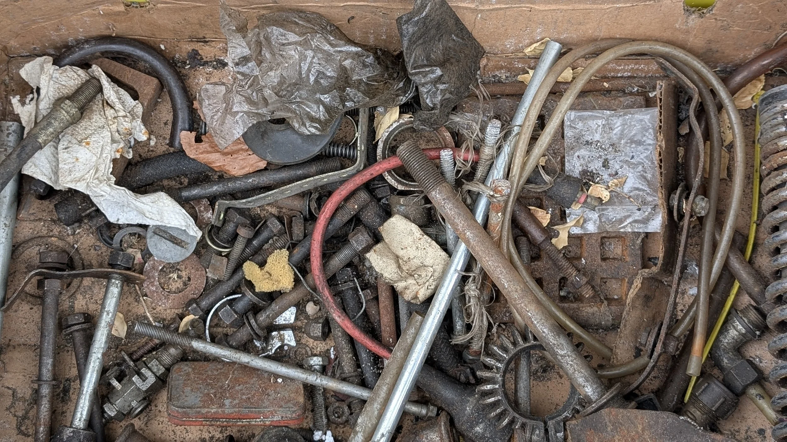 A pile of old and dirty metal bolts, nuts, screws, wires, and scraps on a rust-colored surface. Some crumpled paper and pieces of tape are also visible among the debris. Barn find Triumph T100 restoration project. 