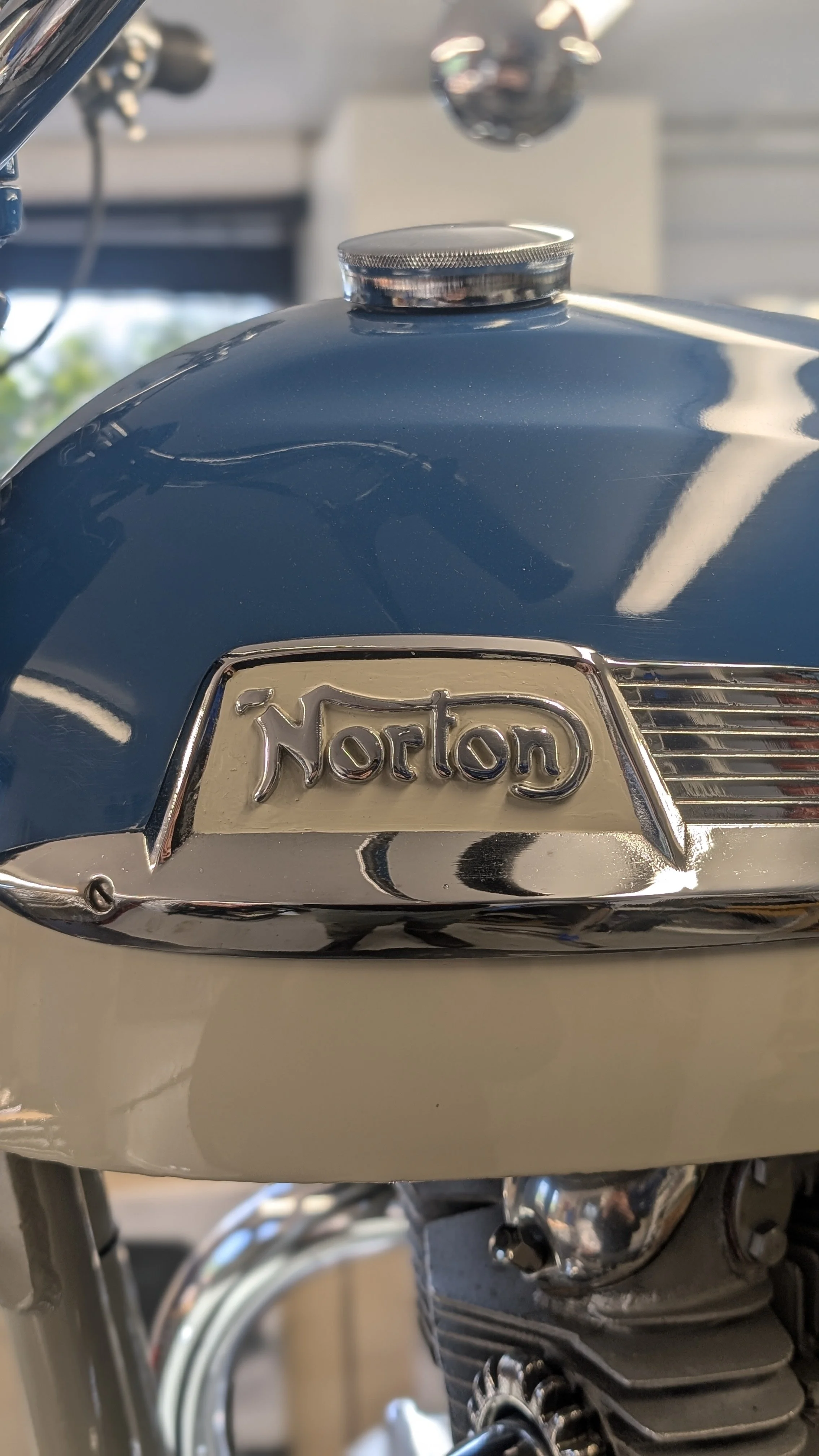 Close-up of a blue motorcycle gas tank with a chrome Norton emblem and fuel cap.