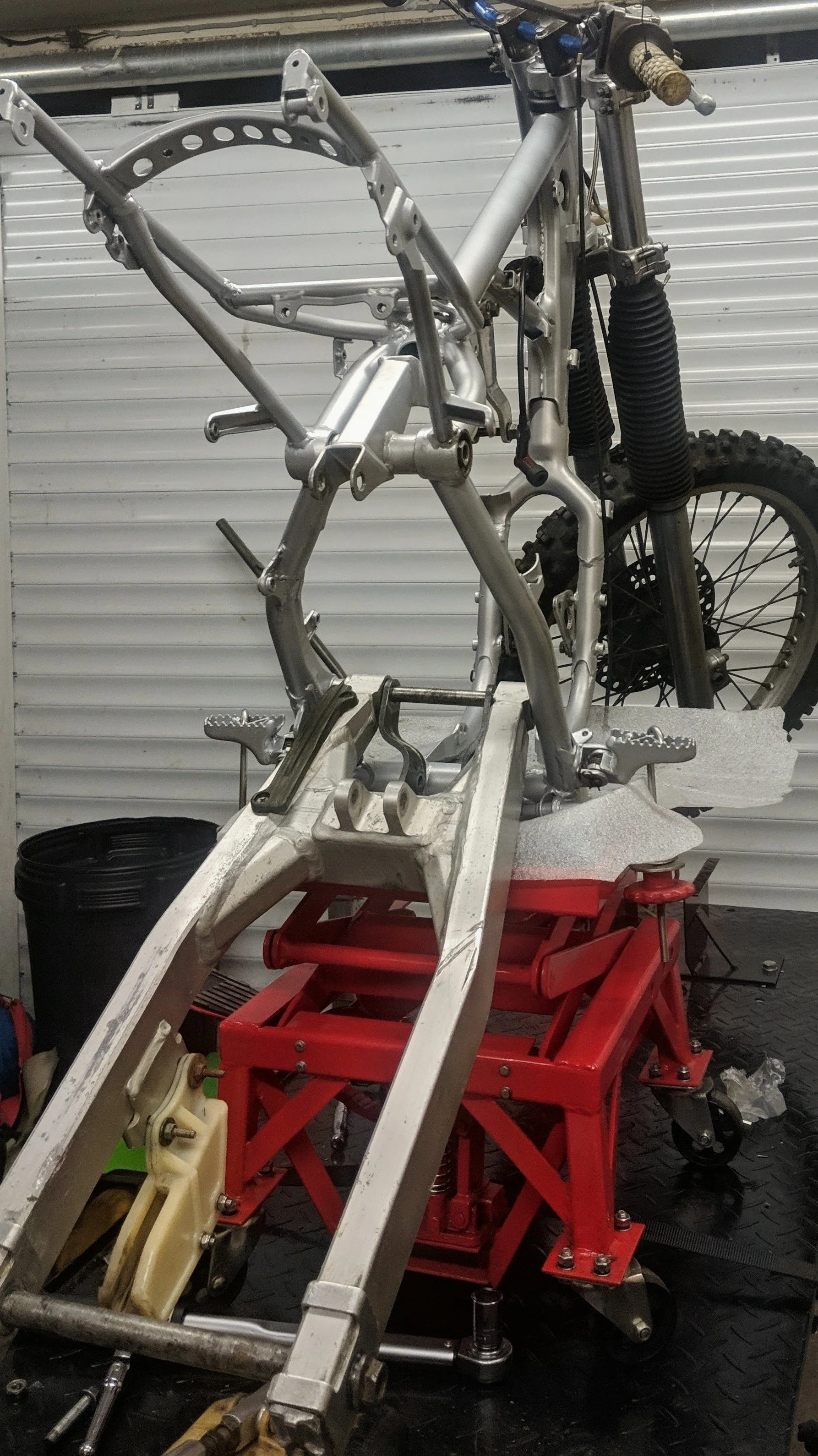 A motorcycle frame under construction, mounted on a work stand in a workshop with tools and equipment around.