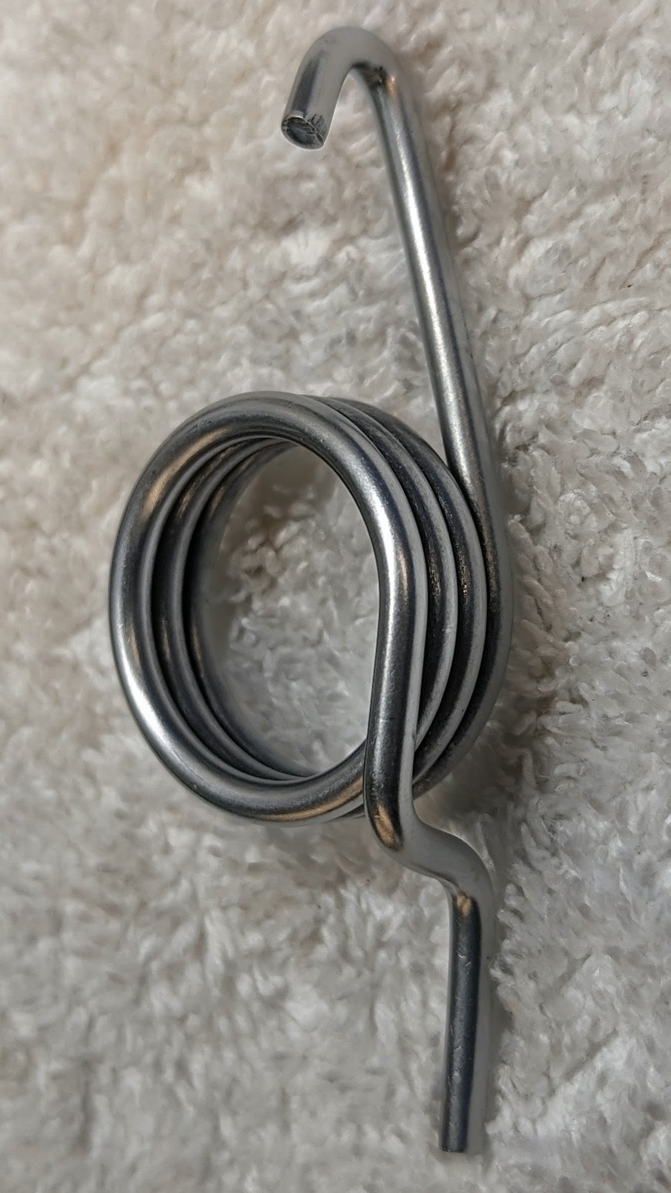 A metal tension spring with a hook at each end is mounted against a textured beige wall.