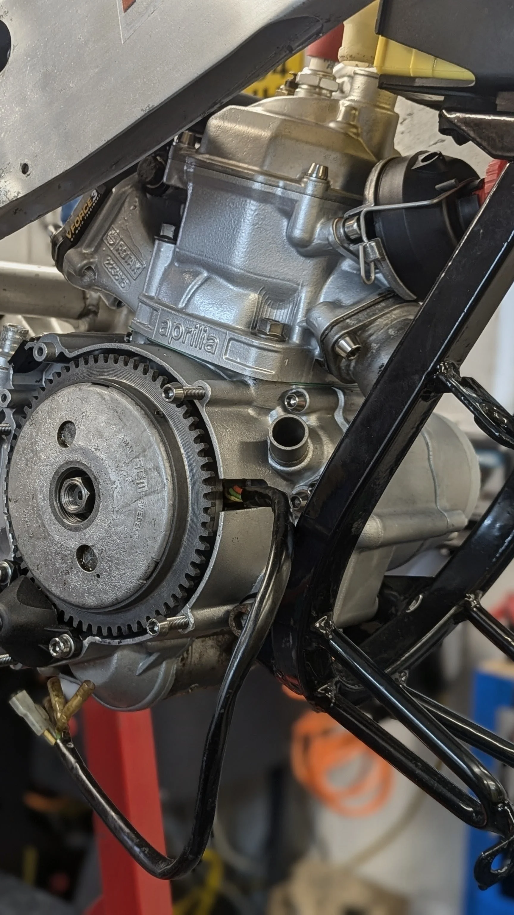Close-up of a motorcycle engine, showing a metallic engine block with the brand name 'aprilia' engraved, a gear attached to the engine, and various connected wires and components.