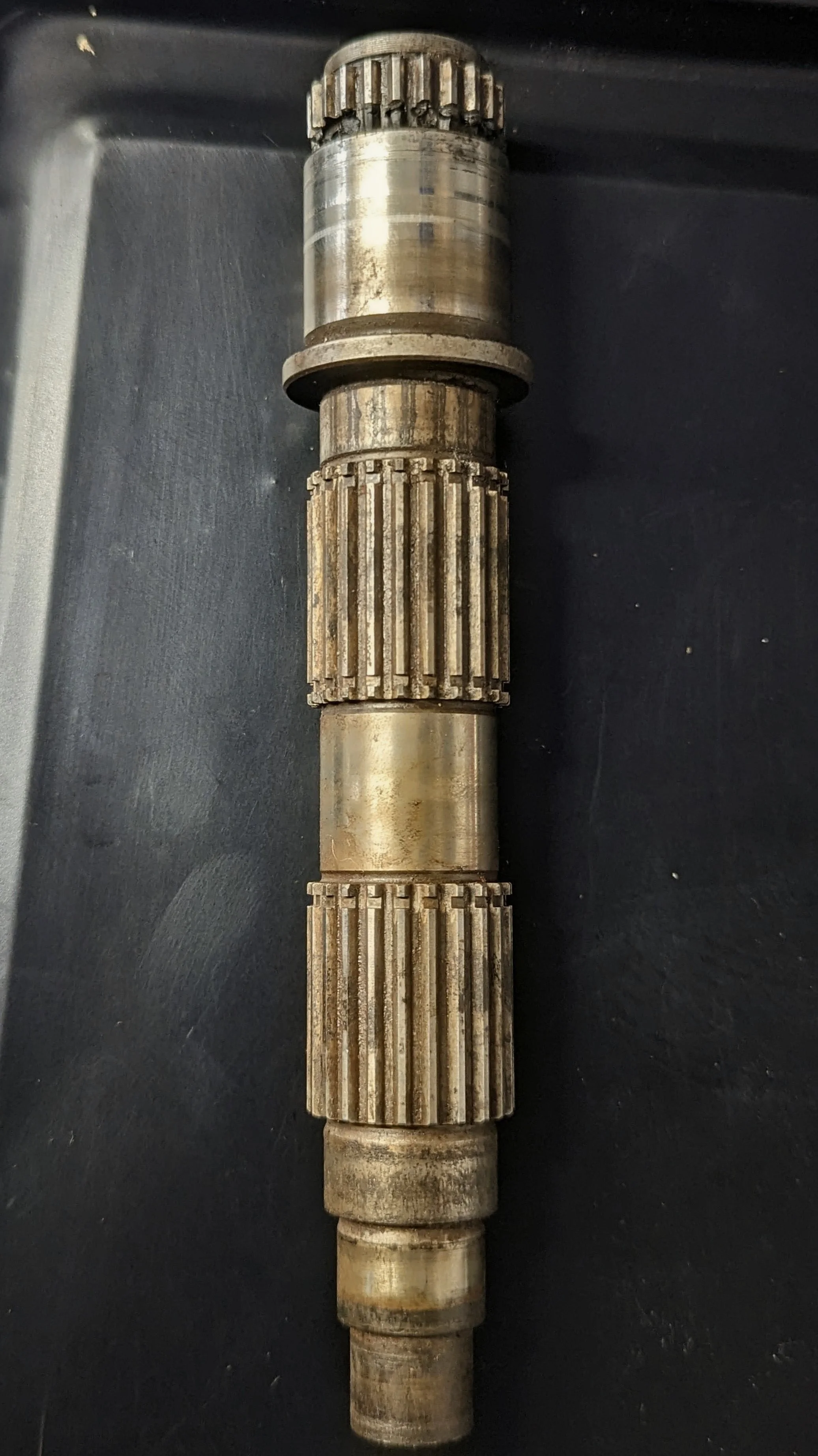 A metal gear shaft with multiple splined sections, some showing signs of rust and wear.