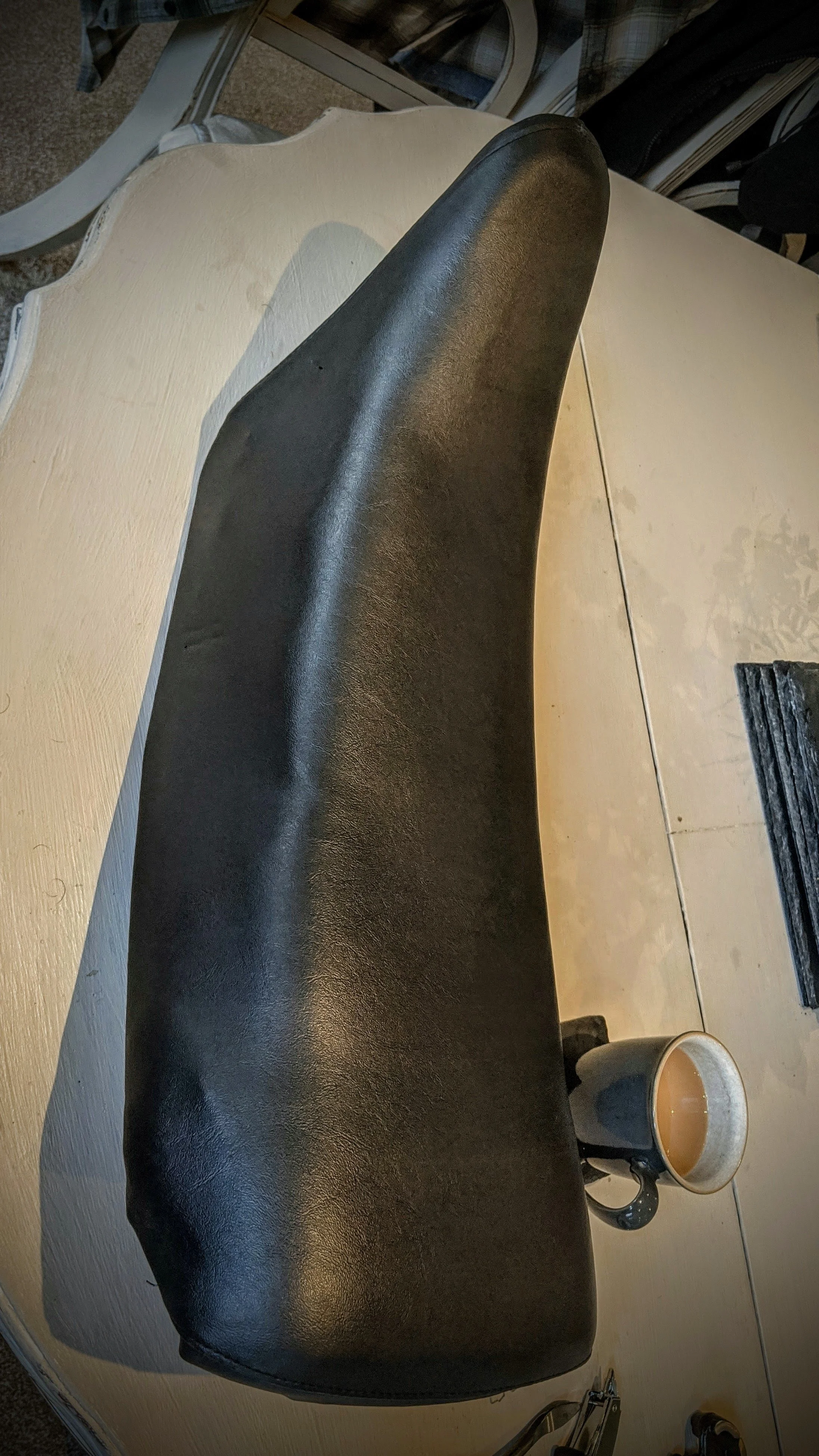 A black leather armrest or headrest extending over a beige wooden table, with a cup of coffee and a cup of tea on the table, surrounded by chairs and other objects.
