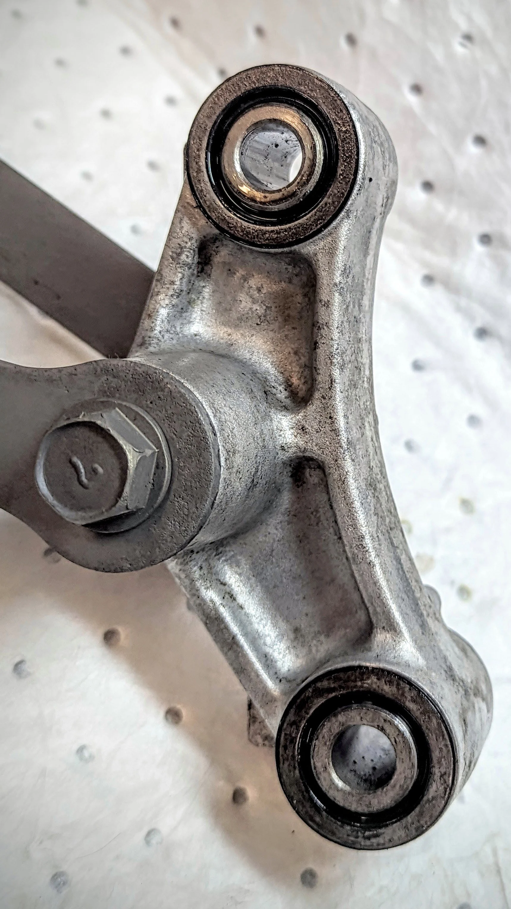 A metal mechanical part with circular bearings and a bolt, resting on a white perforated surface.