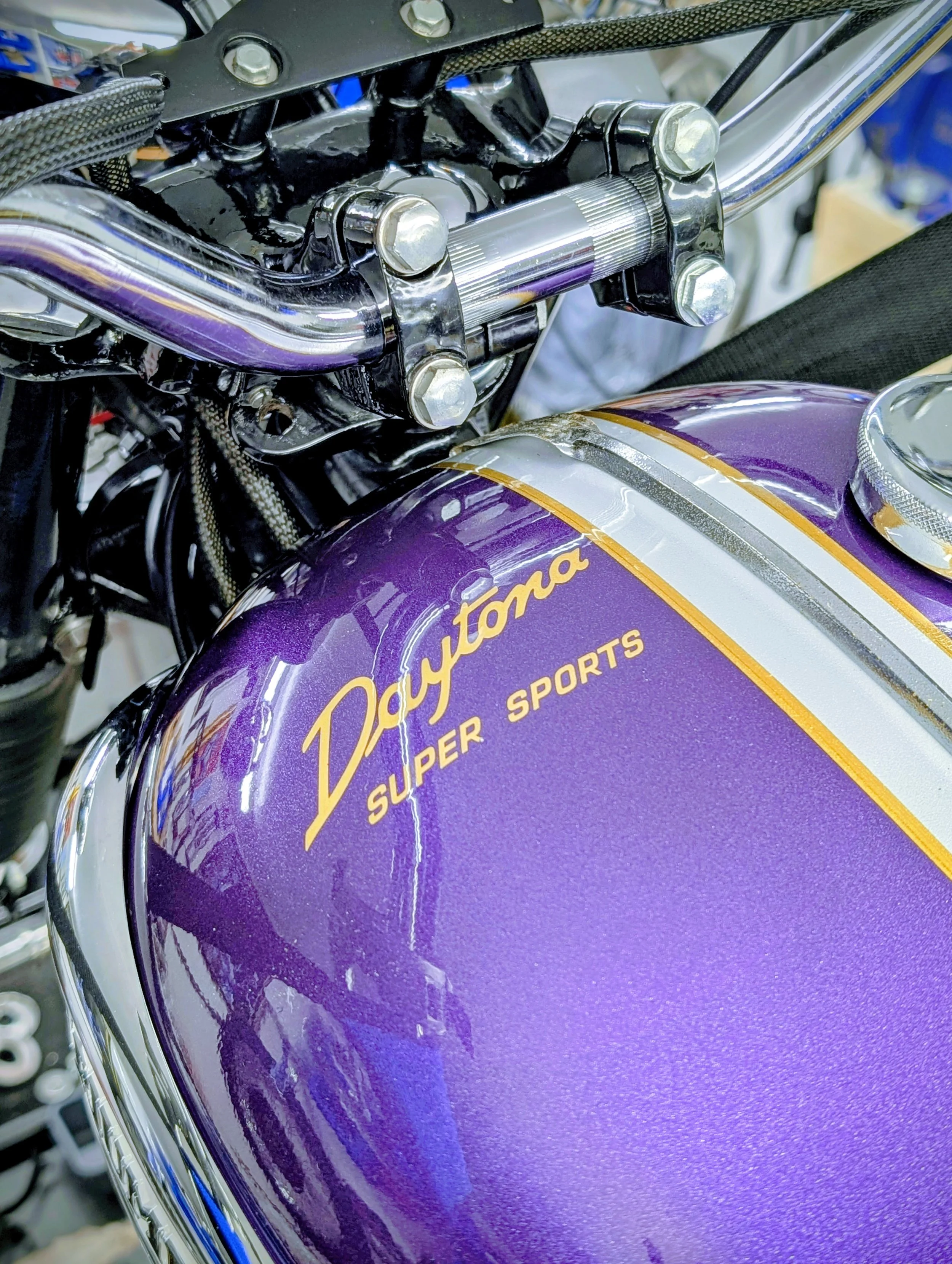 Close-up of a purple motorcycle fuel tank with yellow lettering and chrome accents, showing part of the handlebar and mechanical components. Triumph 500 Daytona T100 fuel tank in original Triumph vintage colours. Barn find Triumph T100 restoration pr