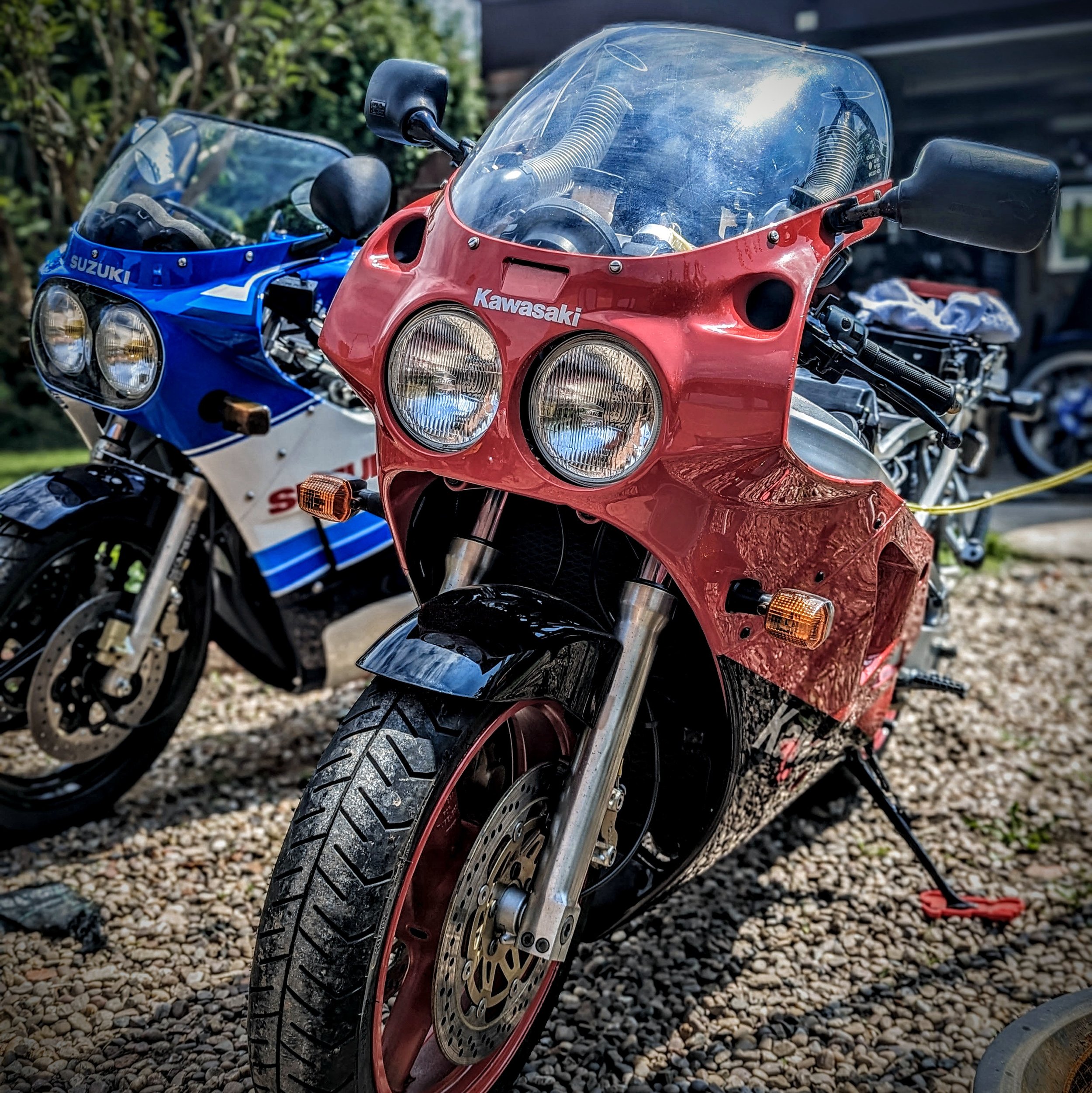 Close-up of a red Kawasaki ZXR sport motorcycle's front end with dual headlights, clear windshield, and front tire, parked outdoors on gravel with other motorcycle, Suzuki GSXR nearby and greenery in the background.