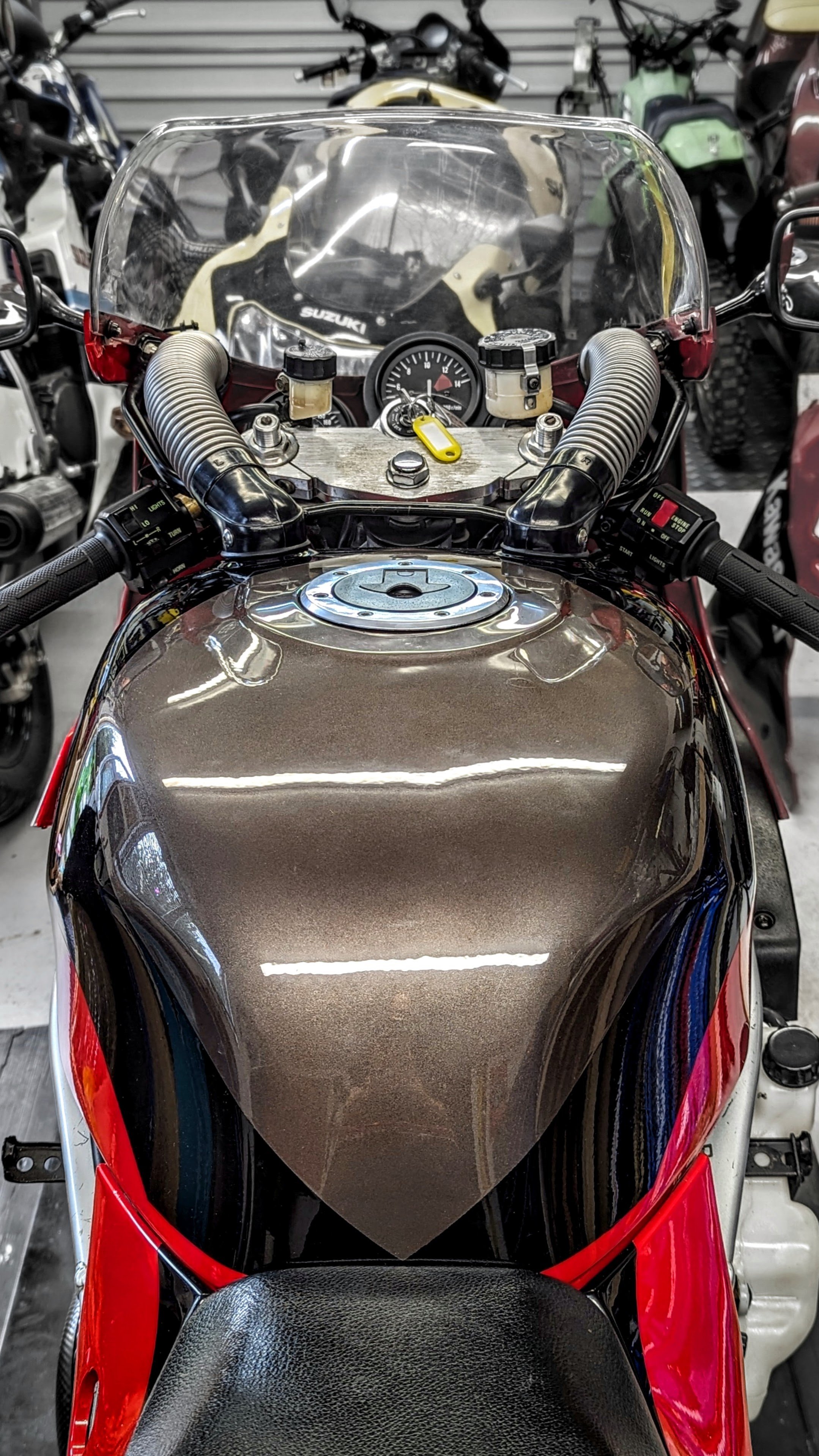 Front view of a motorcycle with a brown metallic fuel tank, black seat, and red fairings, surrounded by other motorcycles in a garage.