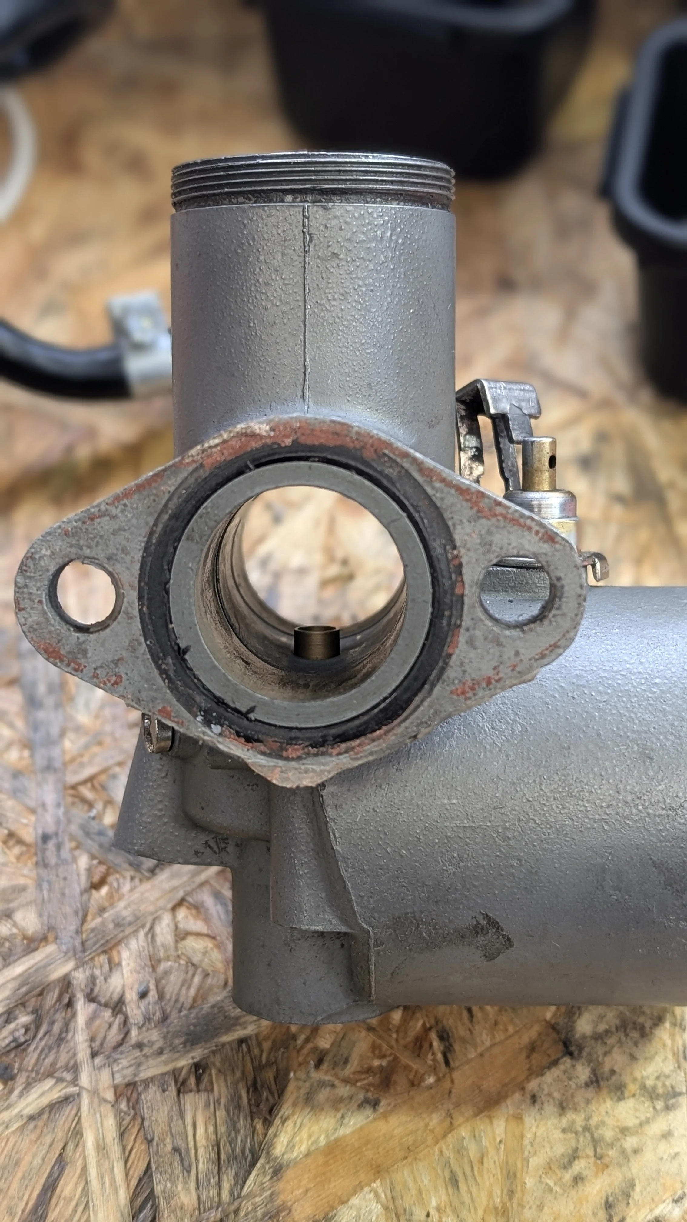A close-up of a gray metal mechanical part with a large central circular opening, some rust around the edges, and a small metal component inside, situated on a wooden surface.
