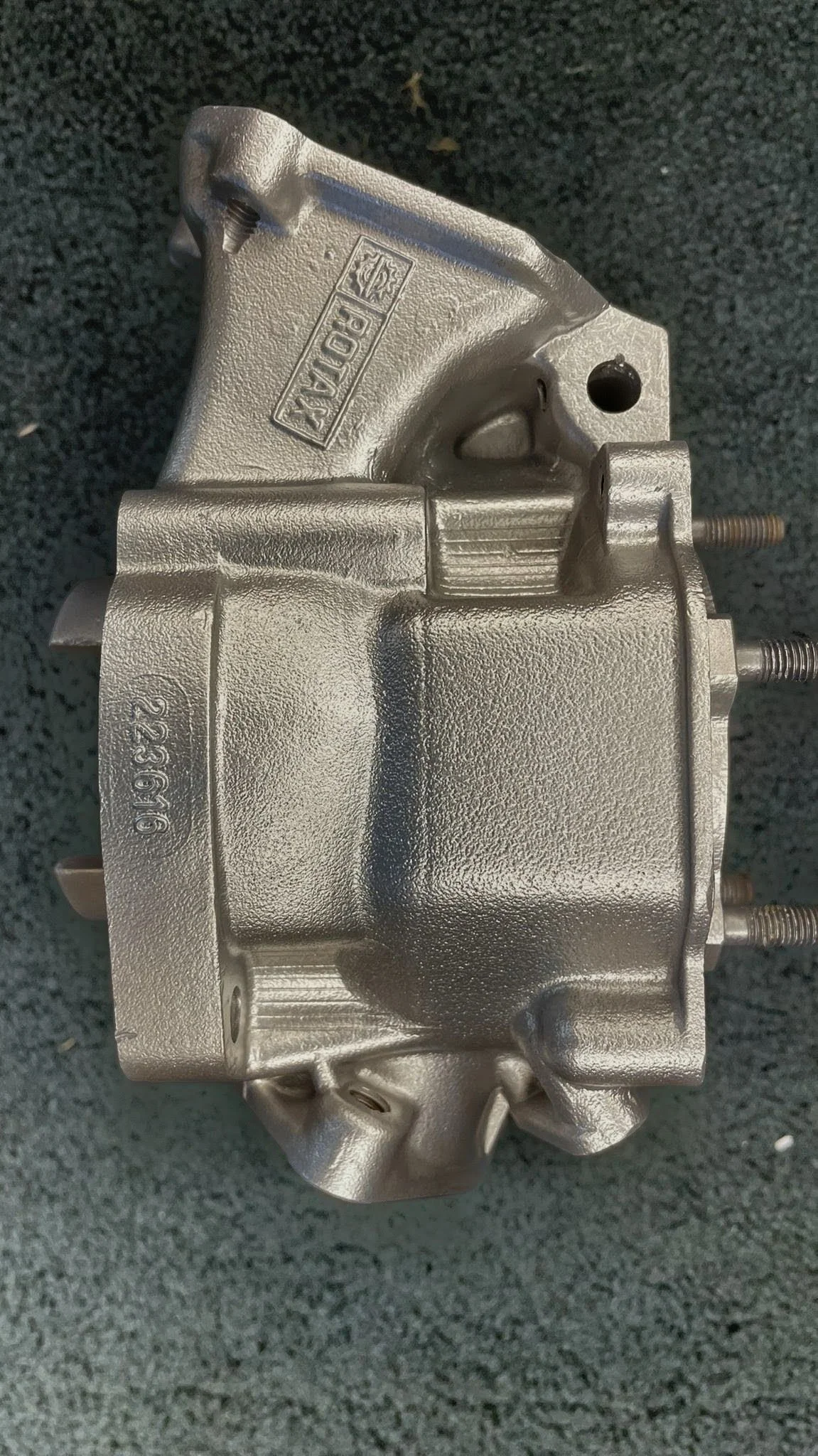 A metallic mechanical component, possibly a part of an engine or machinery, resting on a black textured surface.