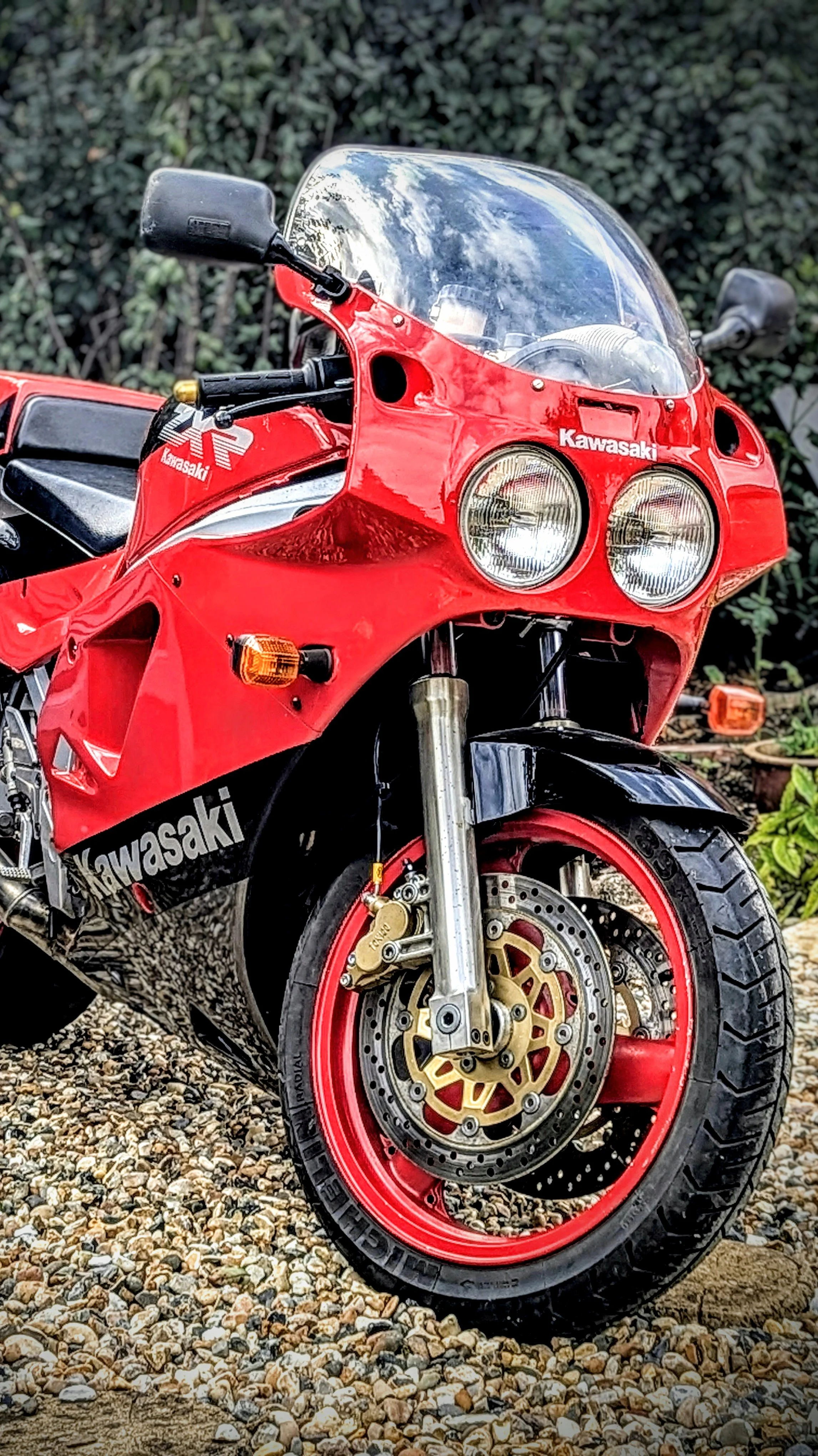 A red Kawasaki motorcycle with a clear windscreen, dual headlights, and a gold front disc brake, parked on gravel with greenery in the background.