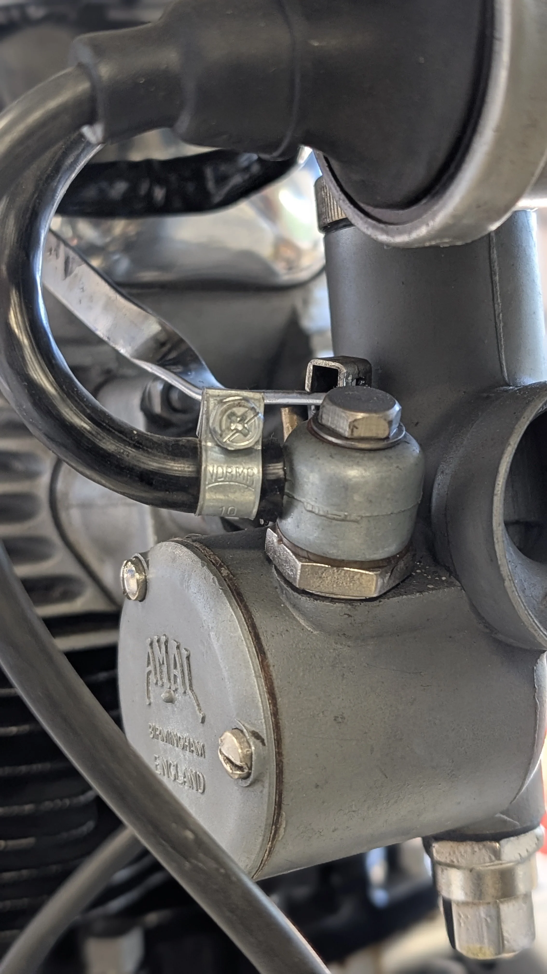 Close-up of a mechanical part on a motorcycle engine, including a metal cylinder with an embossed logo, a brass fitting, a black rubber hose secured with a metal clamp, and various bolts and metal components.