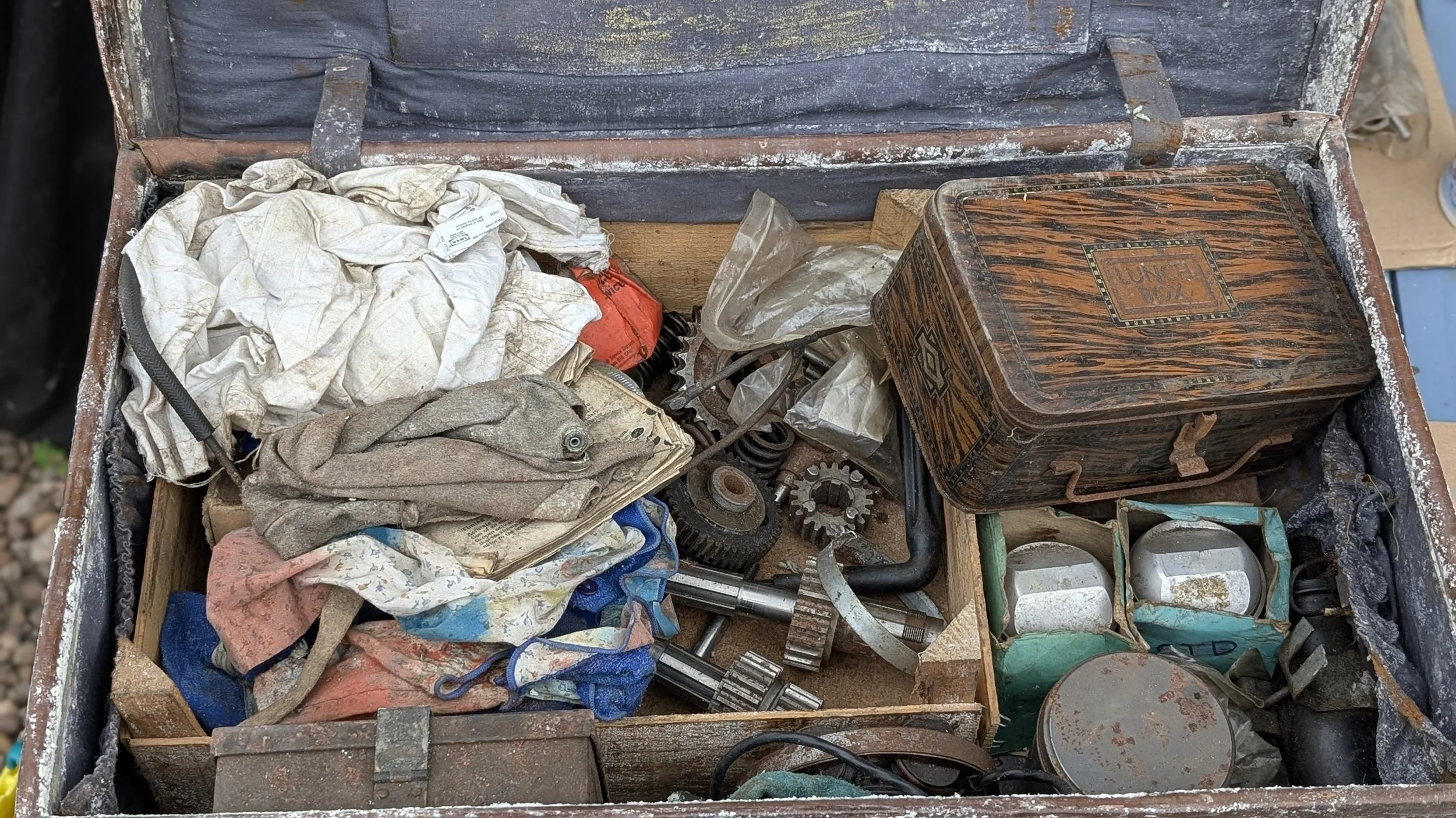 A rusty, open metal toolbox filled with worn rags, mechanical parts, gears, screws, pistons, gearbox gears, sprockets, and miscellaneous tools and objects. Barn find Triumph T100 restoration project. 