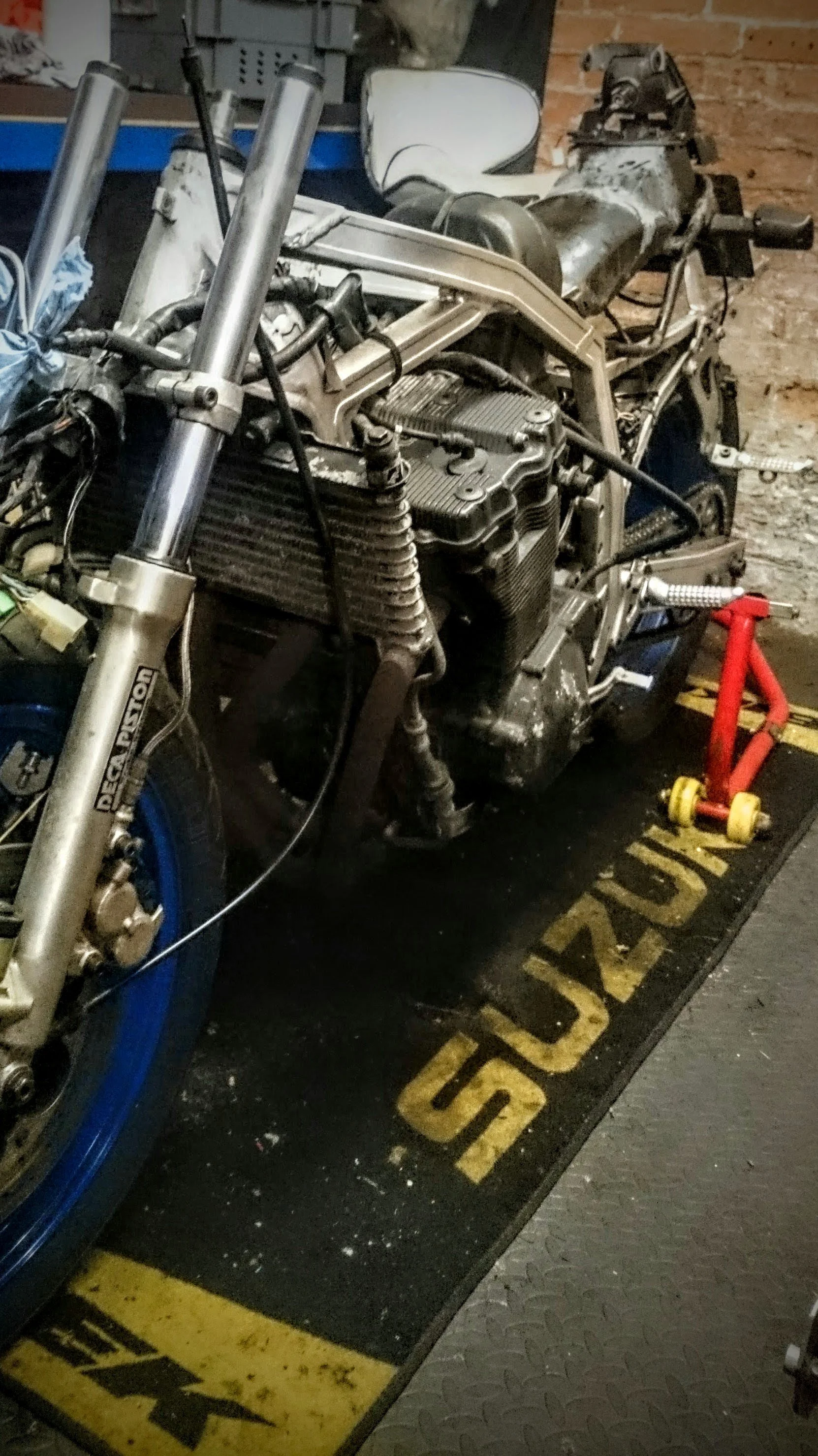 A partially disassembled motorcycle in a workshop with a racing slick mat on the floor. The motorcycle appears to be in the middle of maintenance or restoration, with missing or removed parts and a visible engine.