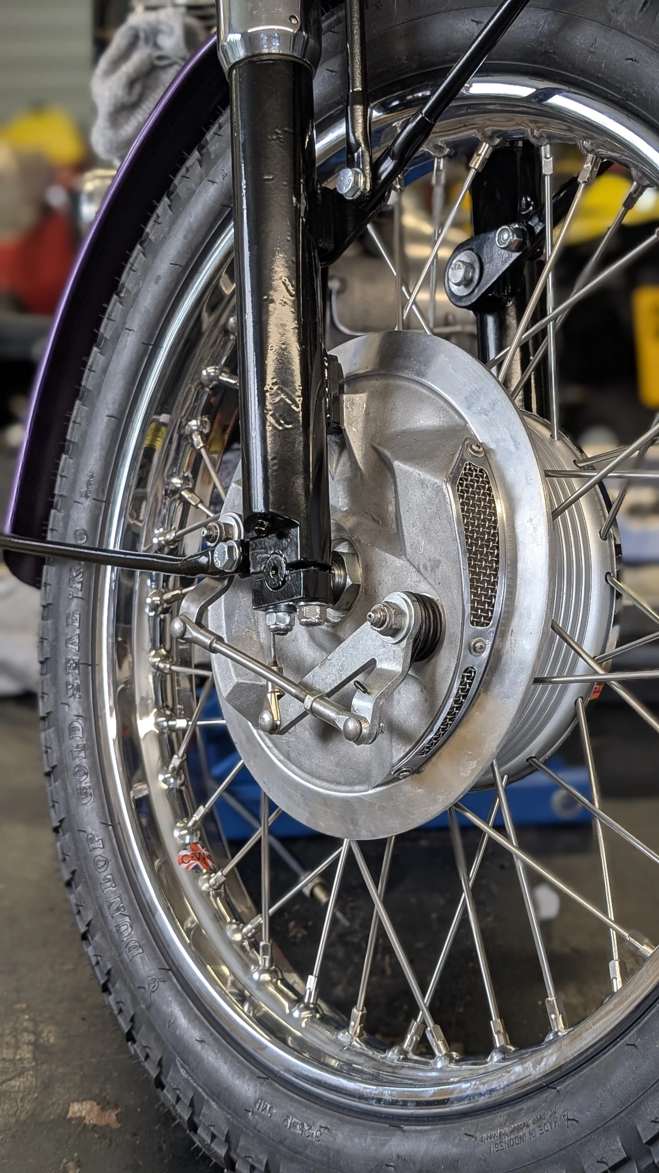 Close-up of a motorcycle wheel, showcasing the TLS twin leading shoe brake, spokes, and part of the suspension. The Dunlop tire has visible treads and markings. The wheel has be restored with English chrome rims and stainless steel spokes as part of 