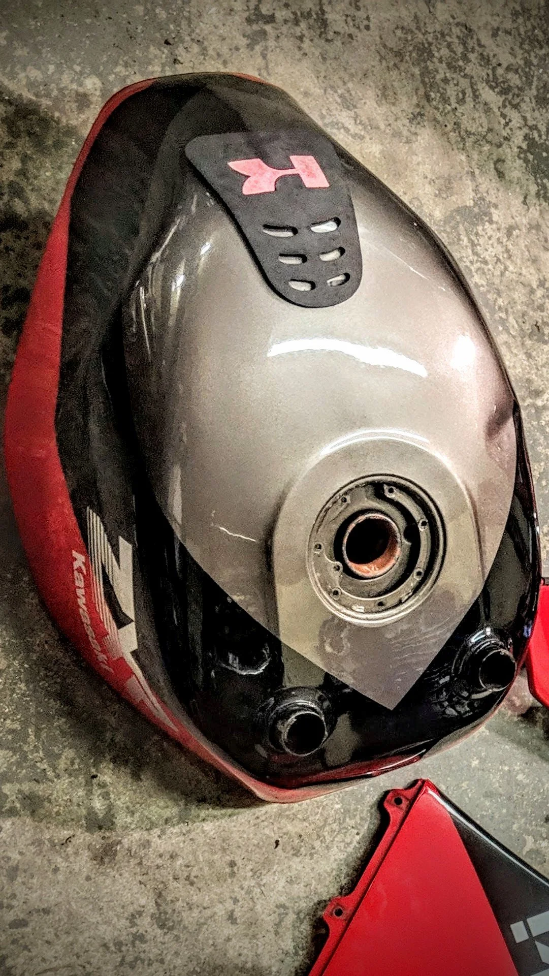 A motorcycle fuel tank removed and placed on a surface, with a black rubber strap on top, and the fuel filler cap area exposed, surrounded by motorcycle parts and tools.