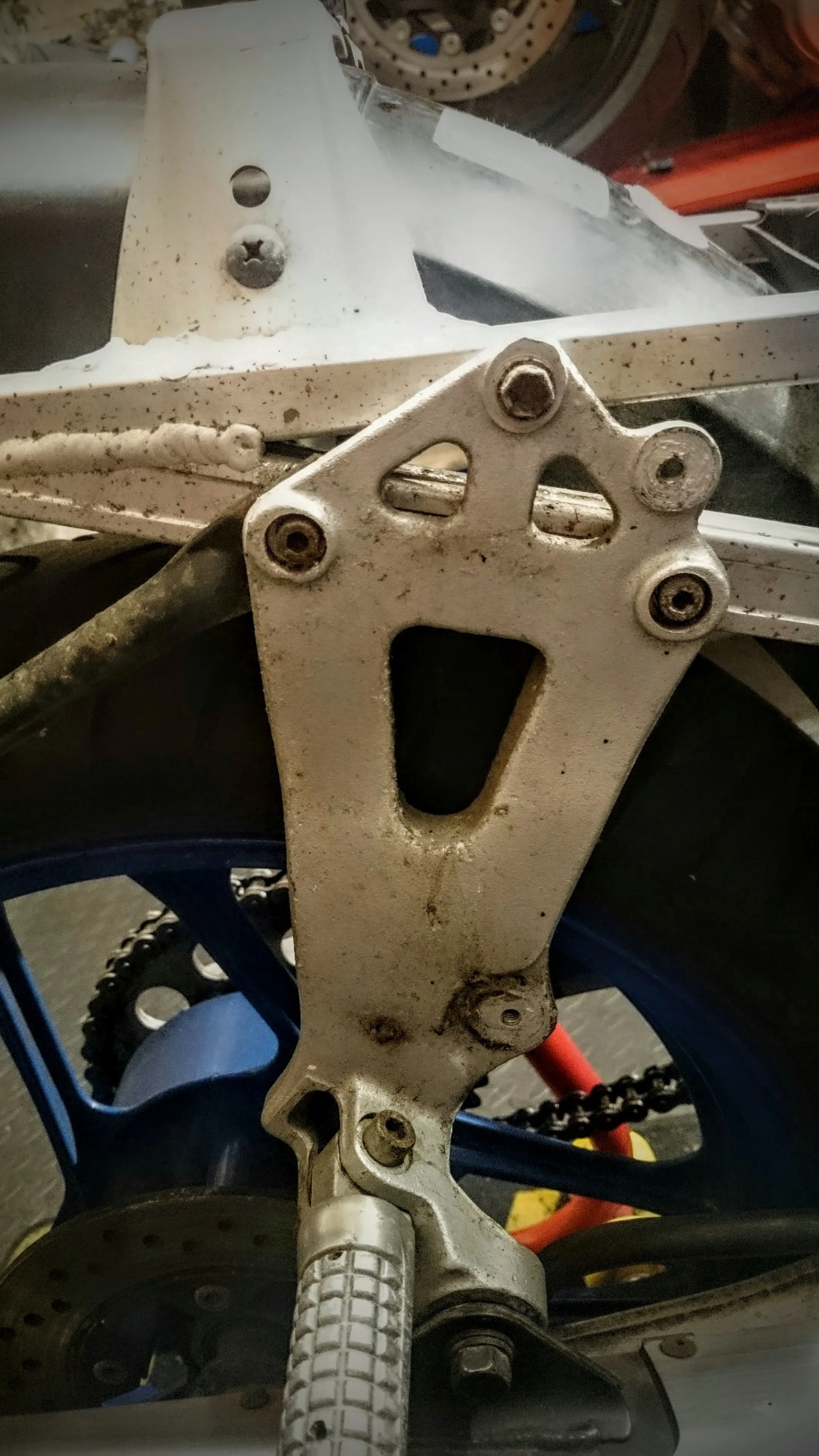 Close-up of a dirt bike frame with a worn white paint job, fastened with screws, showing the chain, chainring, and rear wheel.