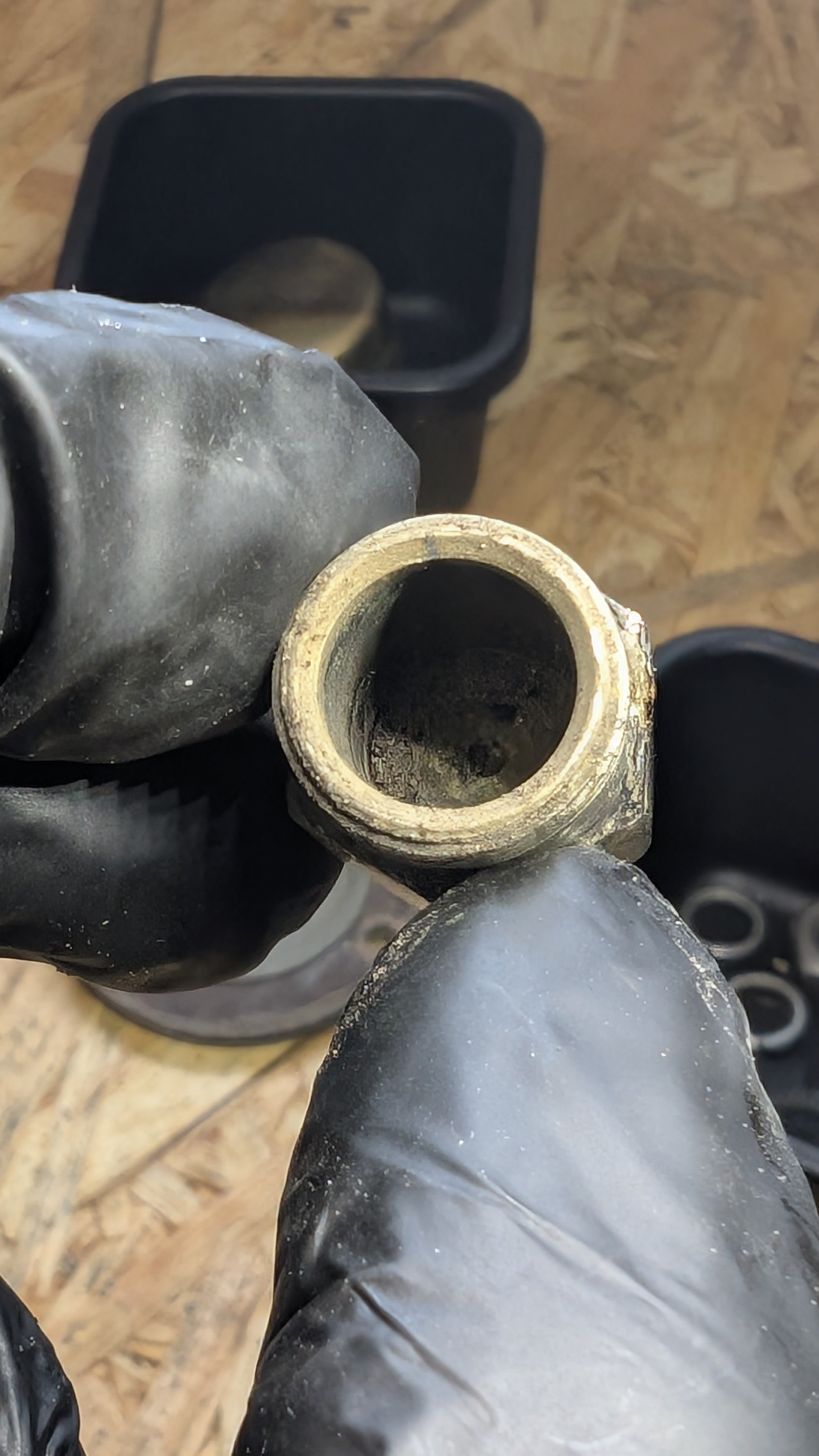Close-up of a person holding a metallic pipe or fitting with a black glove. There's a black container or box and a wooden surface in the background.