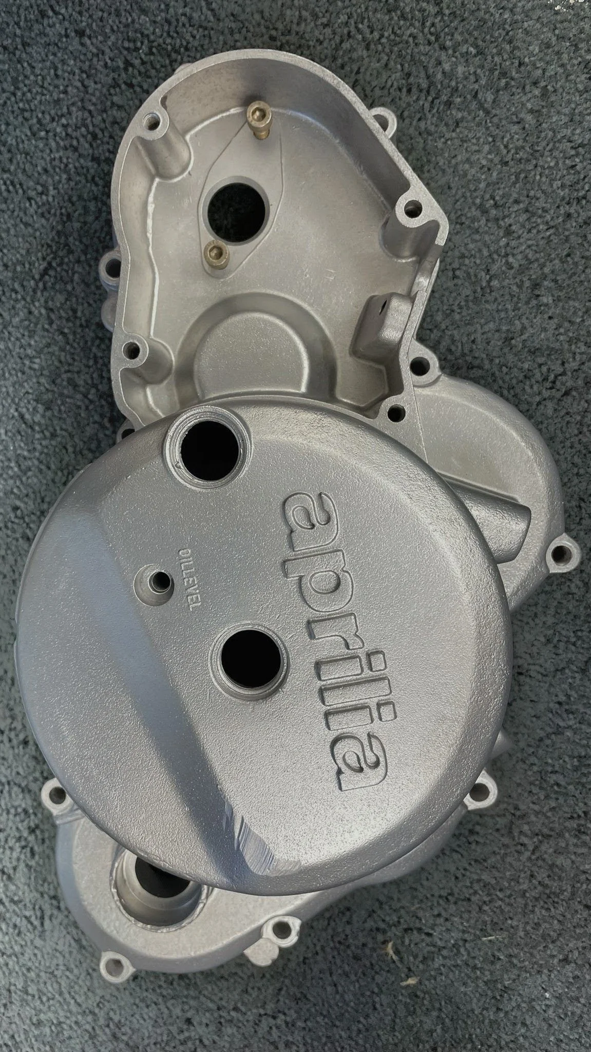 Metal engine cover with the word "aprilia" engraved on it, placed on a gray textured surface.