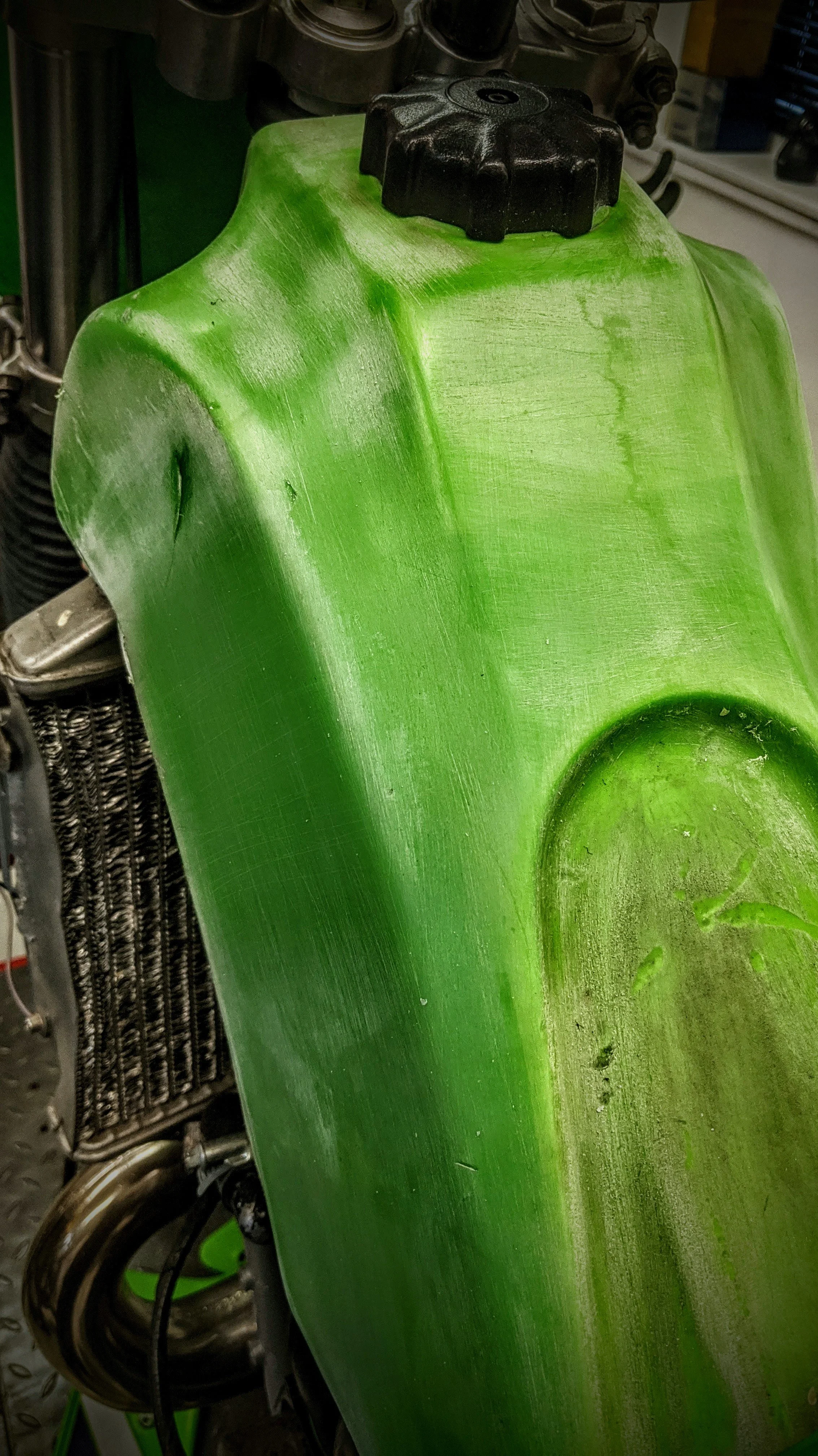 Close-up of a green motorcycle fuel tank with black cap on top, showing scratches and dirt, and parts of the engine and radiator behind it.