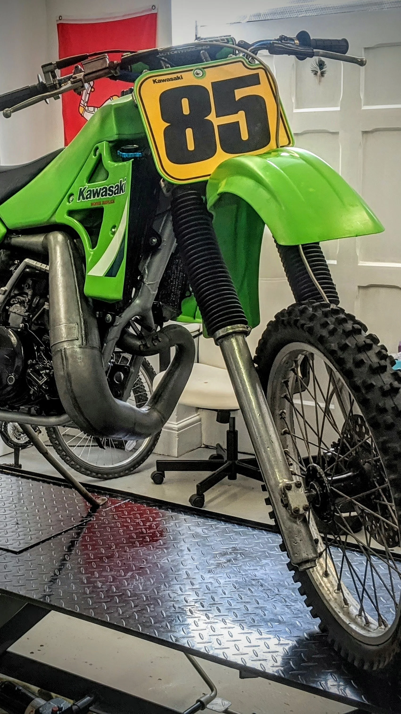 A vintage Kawasaki dirt bike with a yellow number plate labeled 85, green bodywork, and knobby tires, mounted on a metal stand in a room with white walls and a red flag or banner in the background.