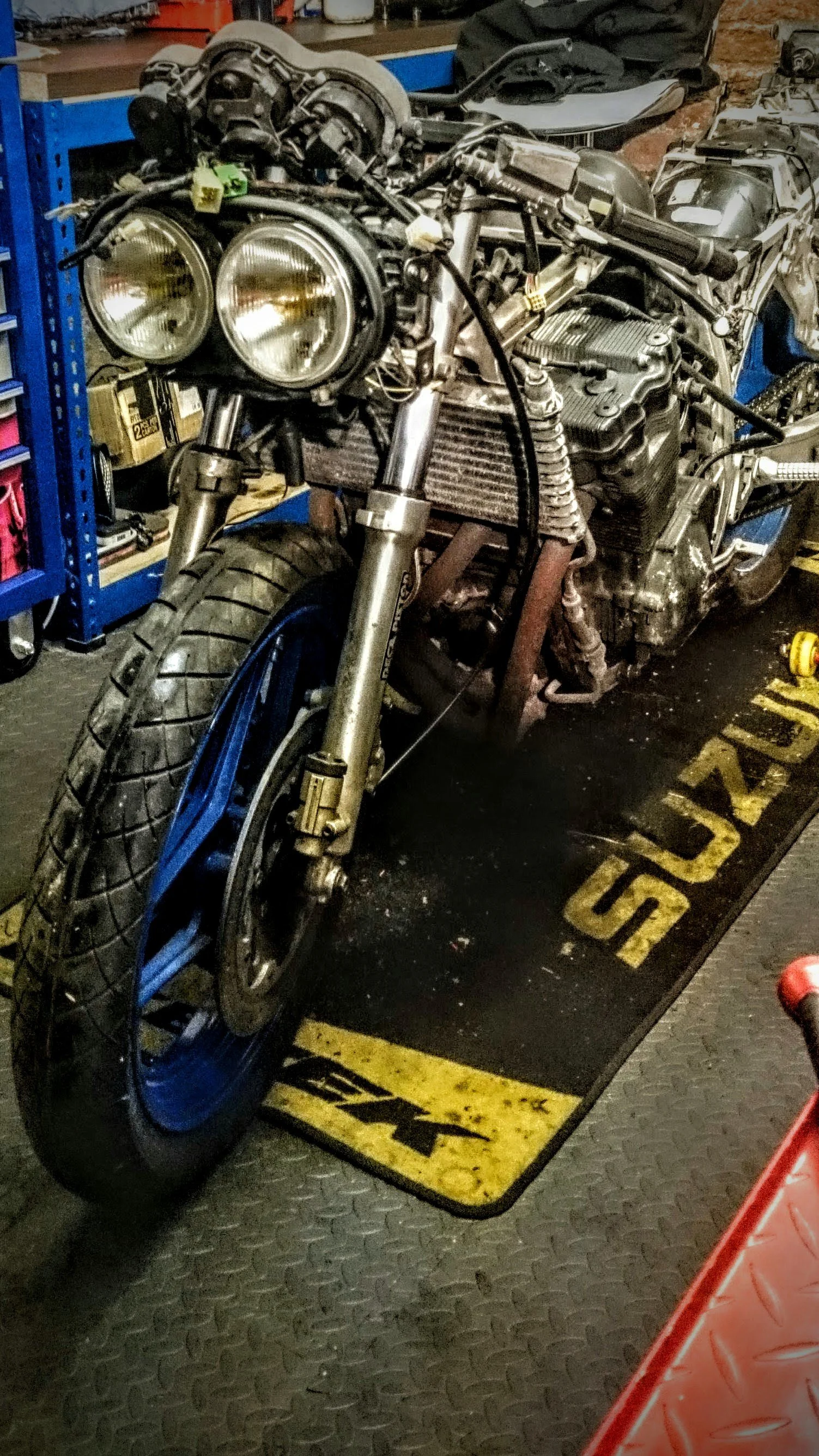 A motorcycle in a workshop with the front tire on a black mat with yellow markings, surrounded by tools and parts. The motorcycle is partially disassembled, exposing the engine and frame.