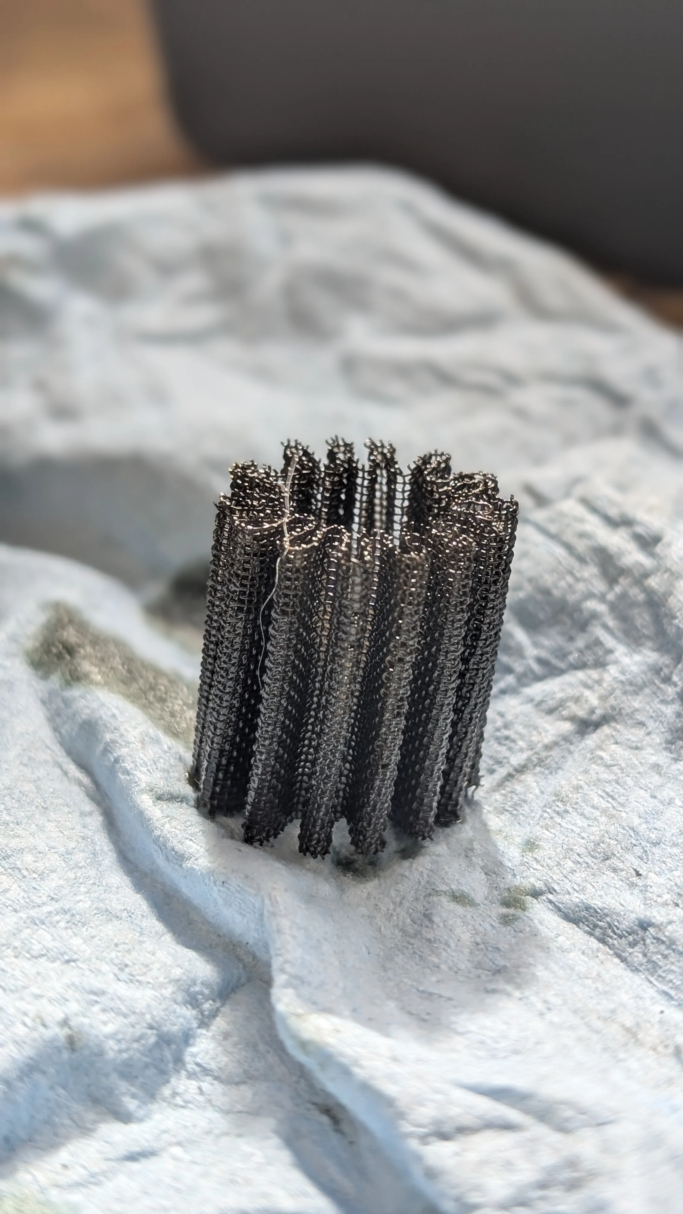 Close-up of a small, metallic, curled-up wire sculpture standing upright on a crumpled white surface.