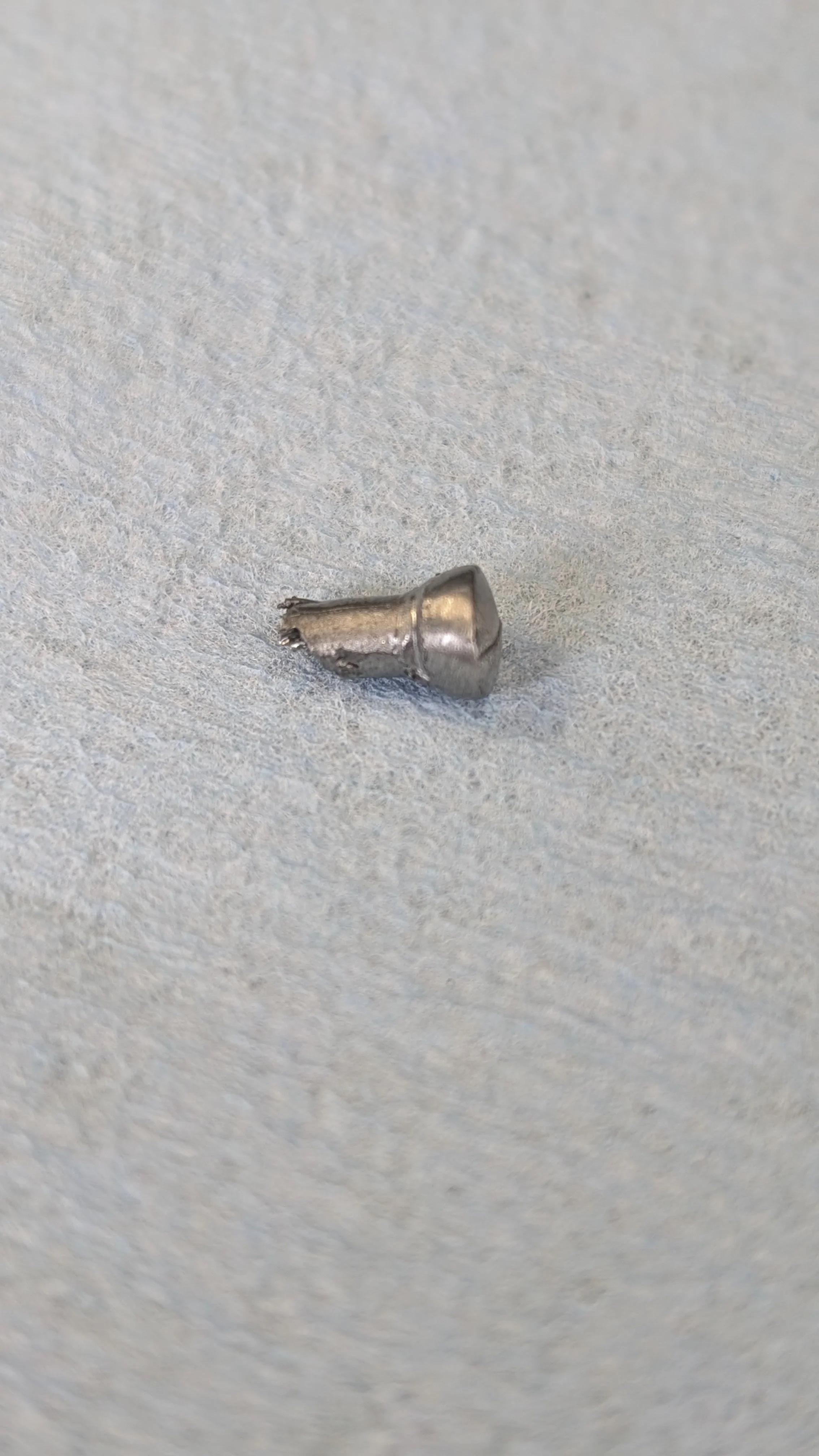 A small, shiny, metallic object resembling a tiny screw or nail on a light gray surface.