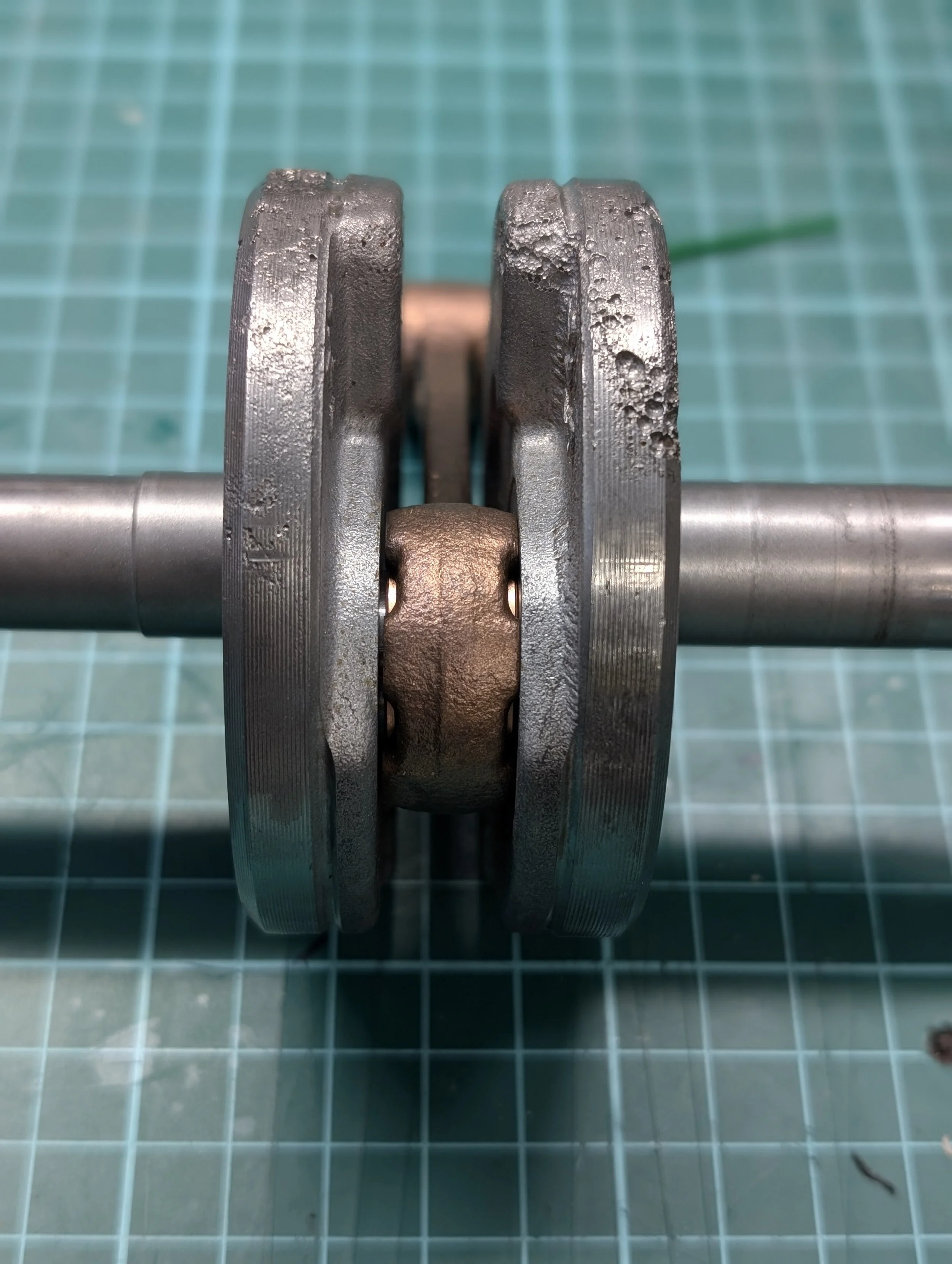 Close-up of a metallic mechanical component with worn edges, placed on a green cutting mat with grid lines, and a thin green tool in the background.
