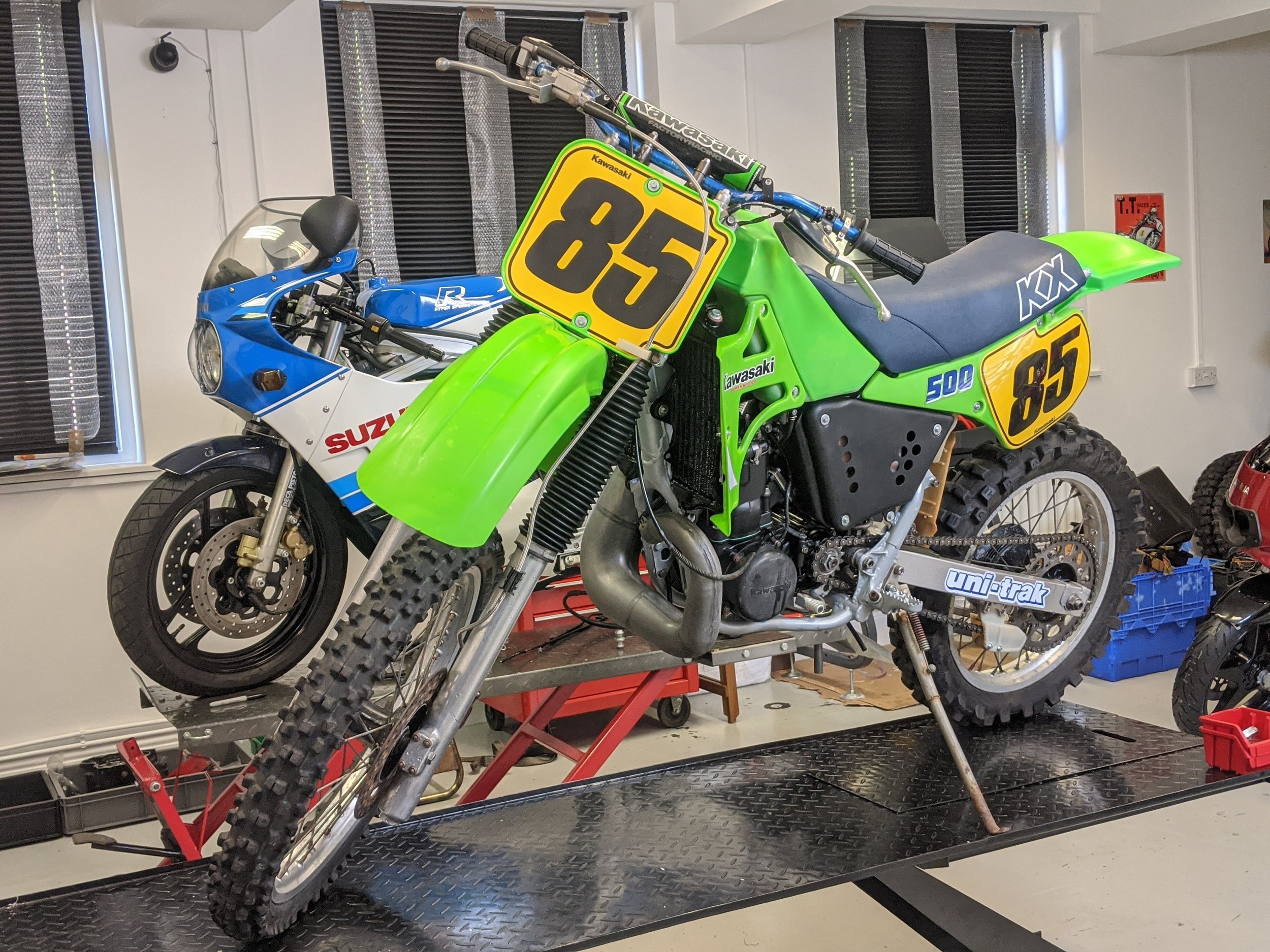 A green and black 1985 Kawasaki KX 500 dirt bike with the number 85 displayed on a yellow plate, mounted on a motorcycle repair or modification stand in a workshop. A 1985 blue and white Suzuki GSX-R 750 slabbie, slabside in the background.