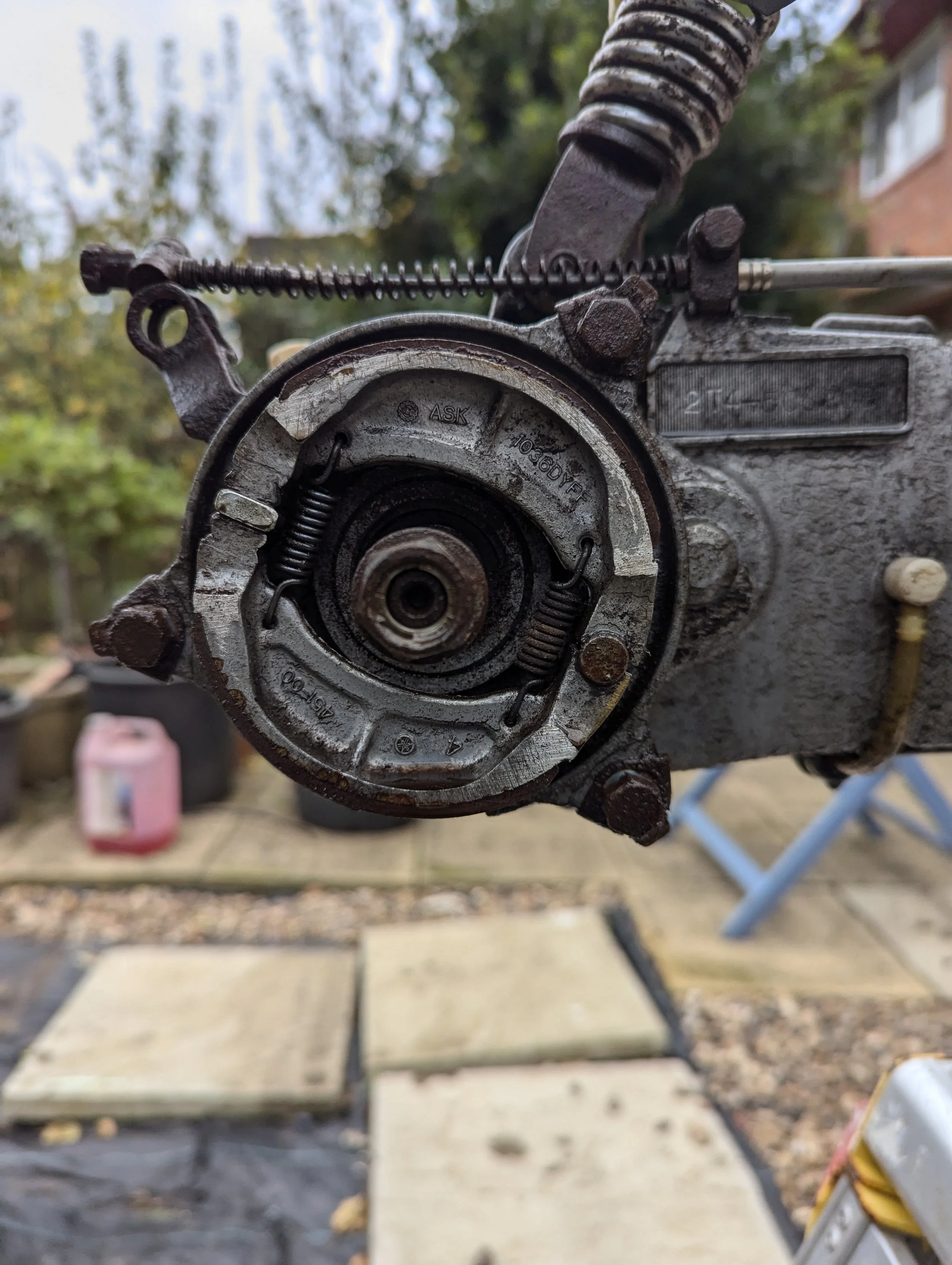 Close-up of a disassembled chainsaw engine part with a spring and metal components, outdoors with trees, a brick house, and a wooden deck in the background.