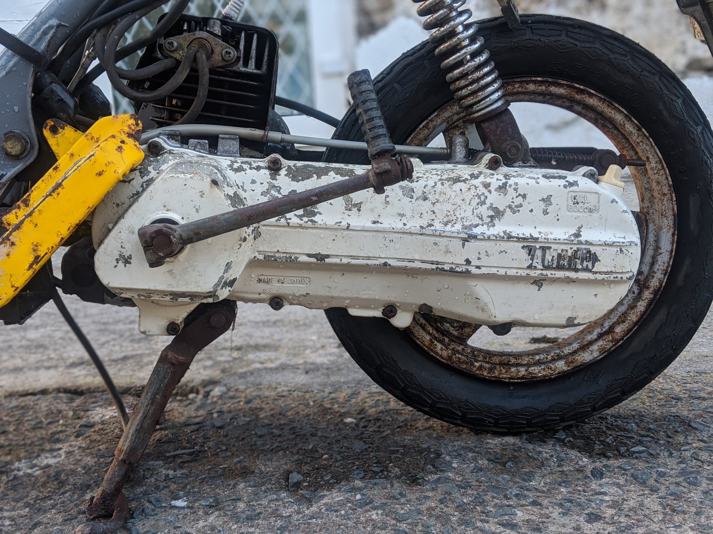 Close-up of an old, rusty motorcycle with a black tire, visible frame, and worn white engine case. The motorcycle appears to be on a gravel surface.