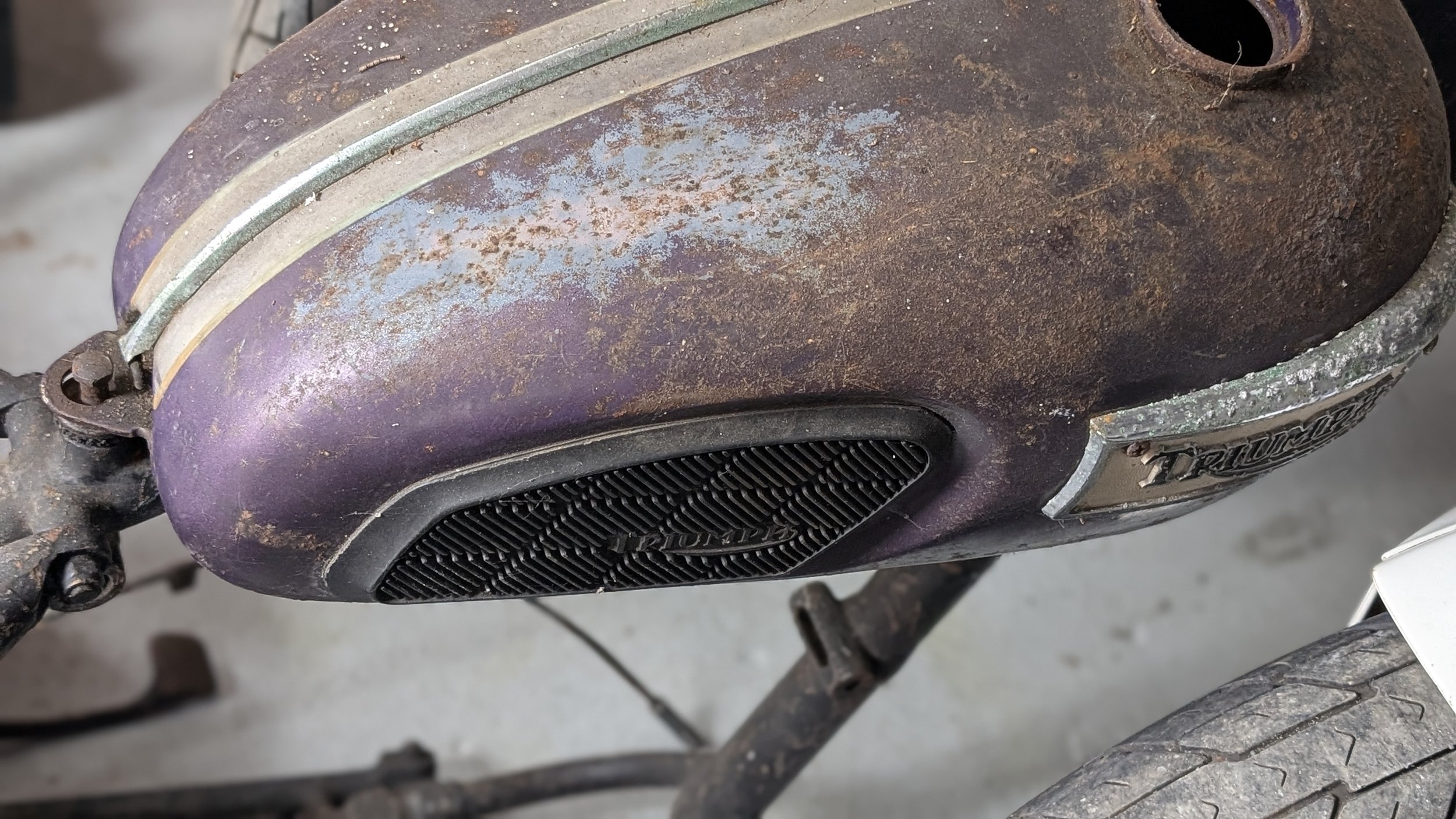 Close-up of a rusty, old motorcycle fuel tank with visible dirt and corrosion. Barn find Triumph T100 restoration project. 