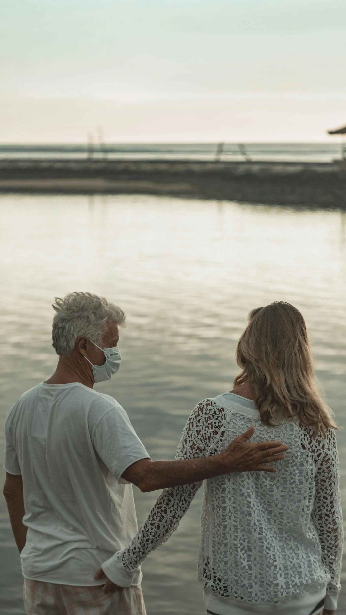 An elderly man and woman stand near a body of water at sunset, with the man wearing a face mask and both holding hands.