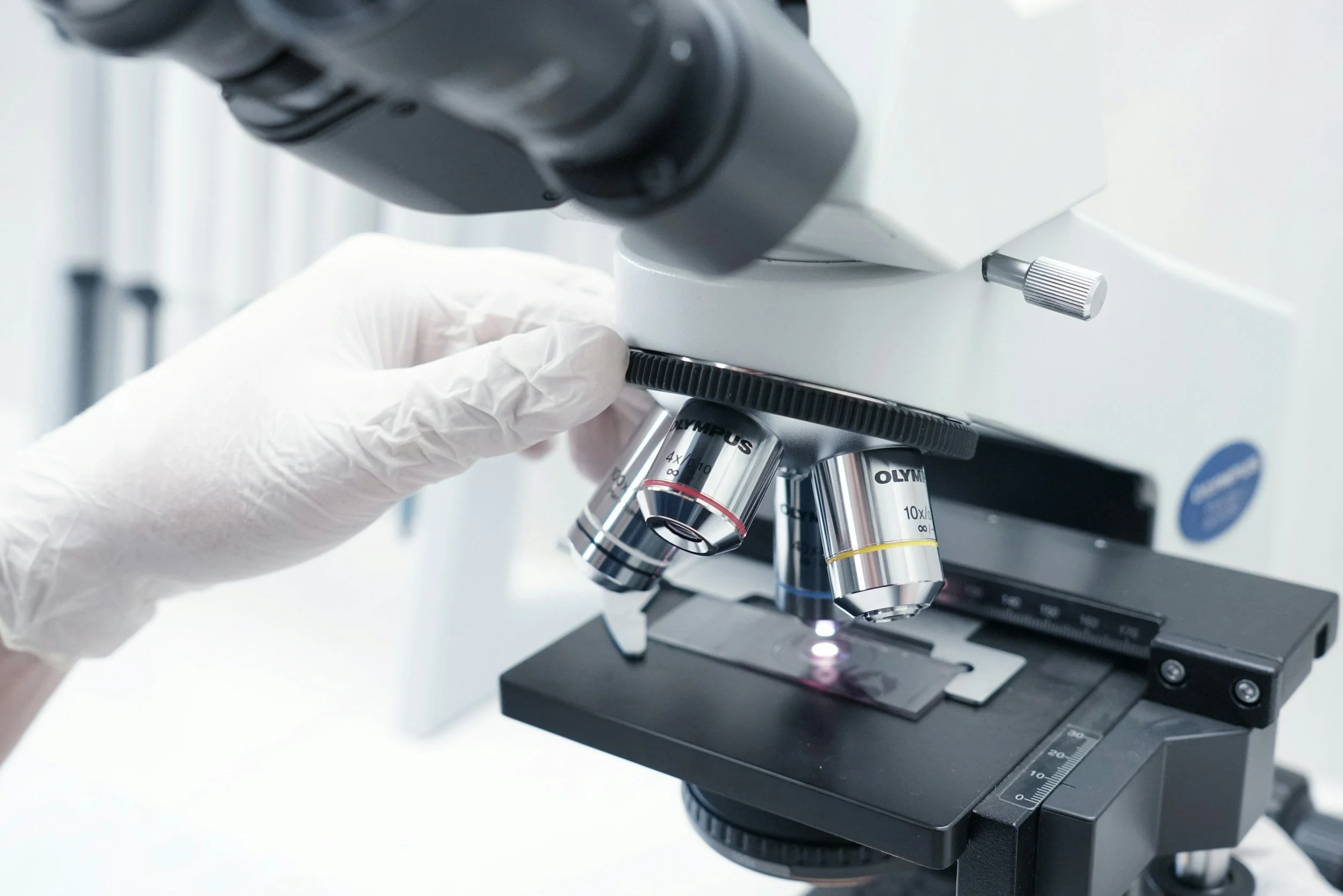 Close-up of a microscope with a gloved hand adjusting the focus knob in a laboratory setting.