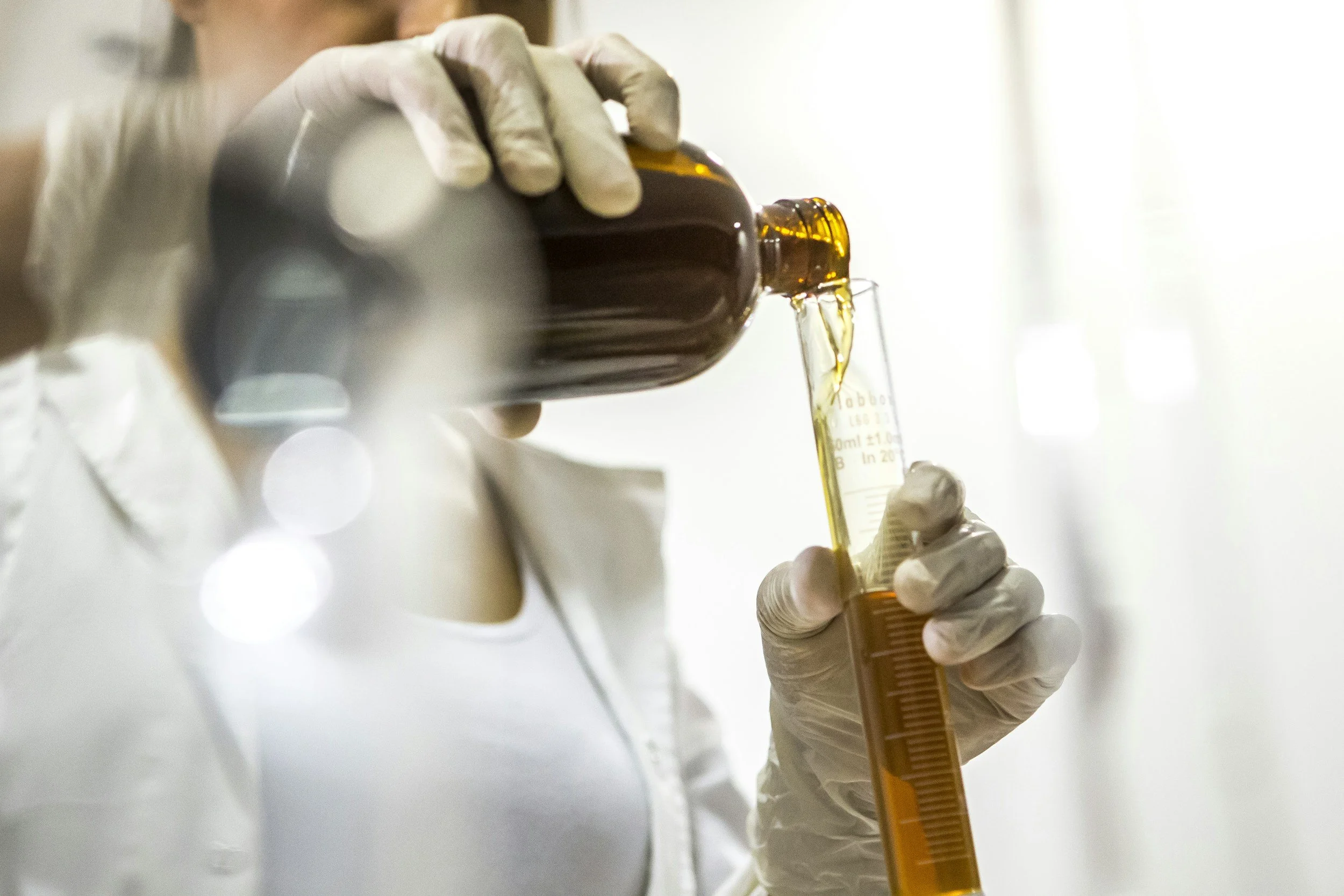 A person wearing white gloves fills a small amber glass bottle with a yellow liquid using a syringe or pipette, with a blurred background.