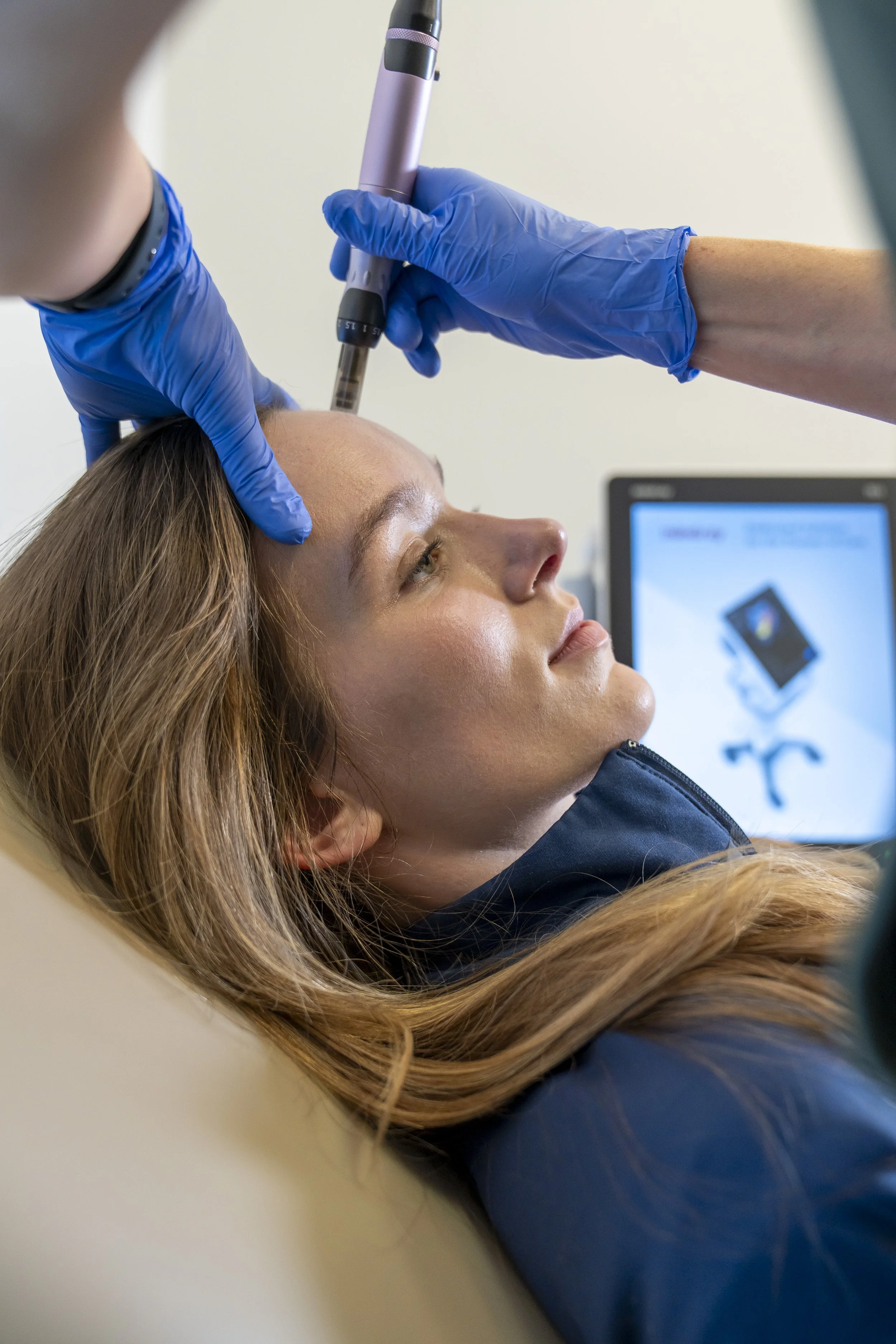 A woman with long, wavy red hair is lying down with her head resting on a surface. Someone wearing blue gloves is holding an ultrasound probe against her forehead. A monitor with a display is visible in the background.
