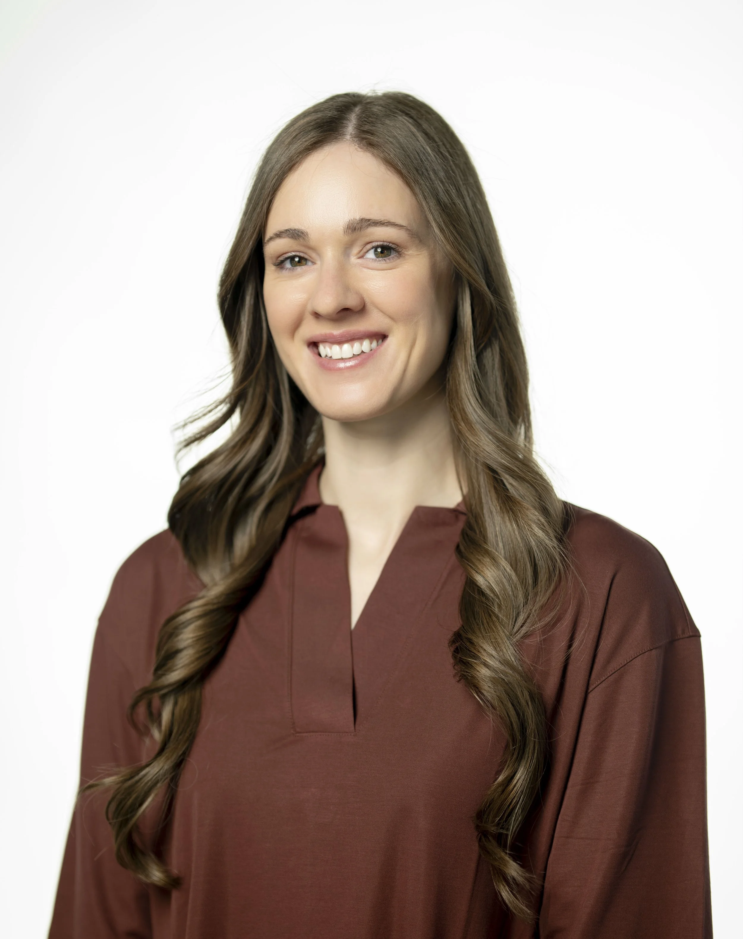 A young woman with long, wavy brown hair and green eyes smiling, wearing a brown long-sleeve top, against a plain white background.