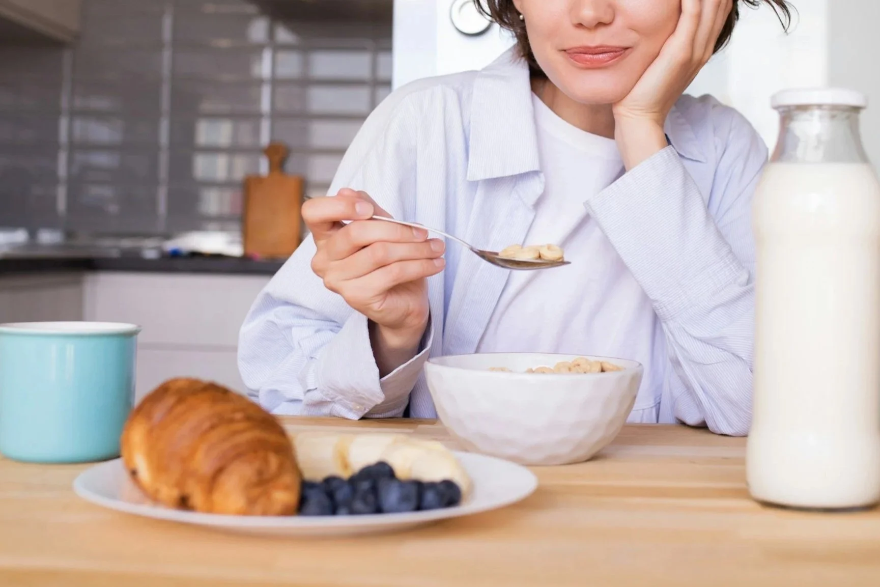 A woman in a white shirt sitting at a kitchen counter, holding a spoon with cereal, smiling lightly. There is cereal in a white bowl, a croissant, blueberries, and a bottle of milk on the counter, with a teal mug also visible.