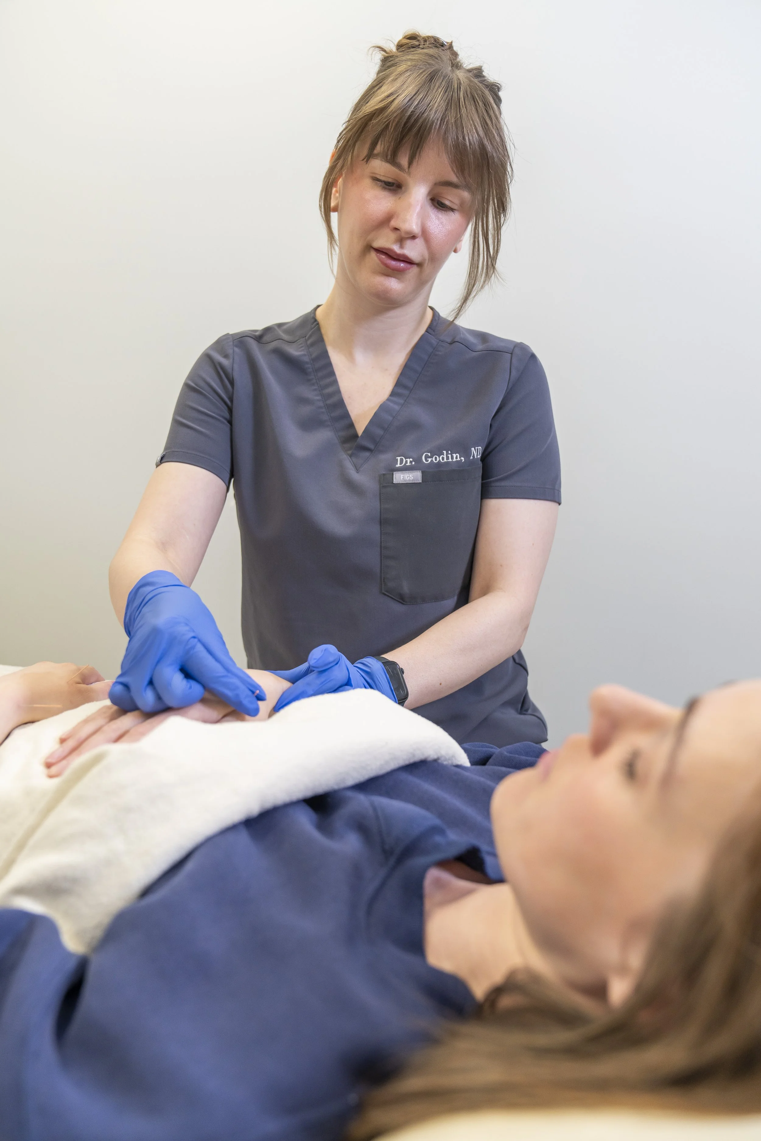 A healthcare professional, wearing gray scrubs and blue gloves, is examining a patient's hand. The patient is lying down on a bed with a white towel over their chest, and appears relaxed. The healthcare professional is focused on the examination, and the background is plain white.