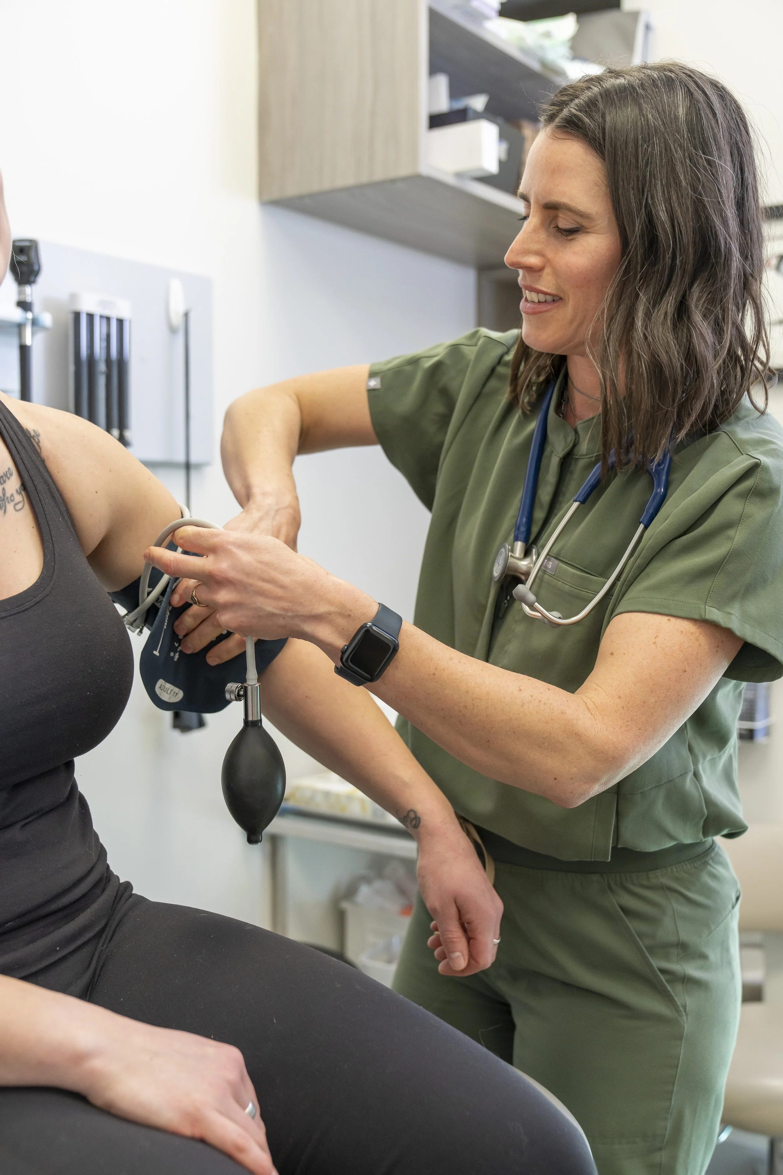 A healthcare professional in green scrubs and a stethoscope taking a patient's blood pressure with a manual blood pressure cuff.