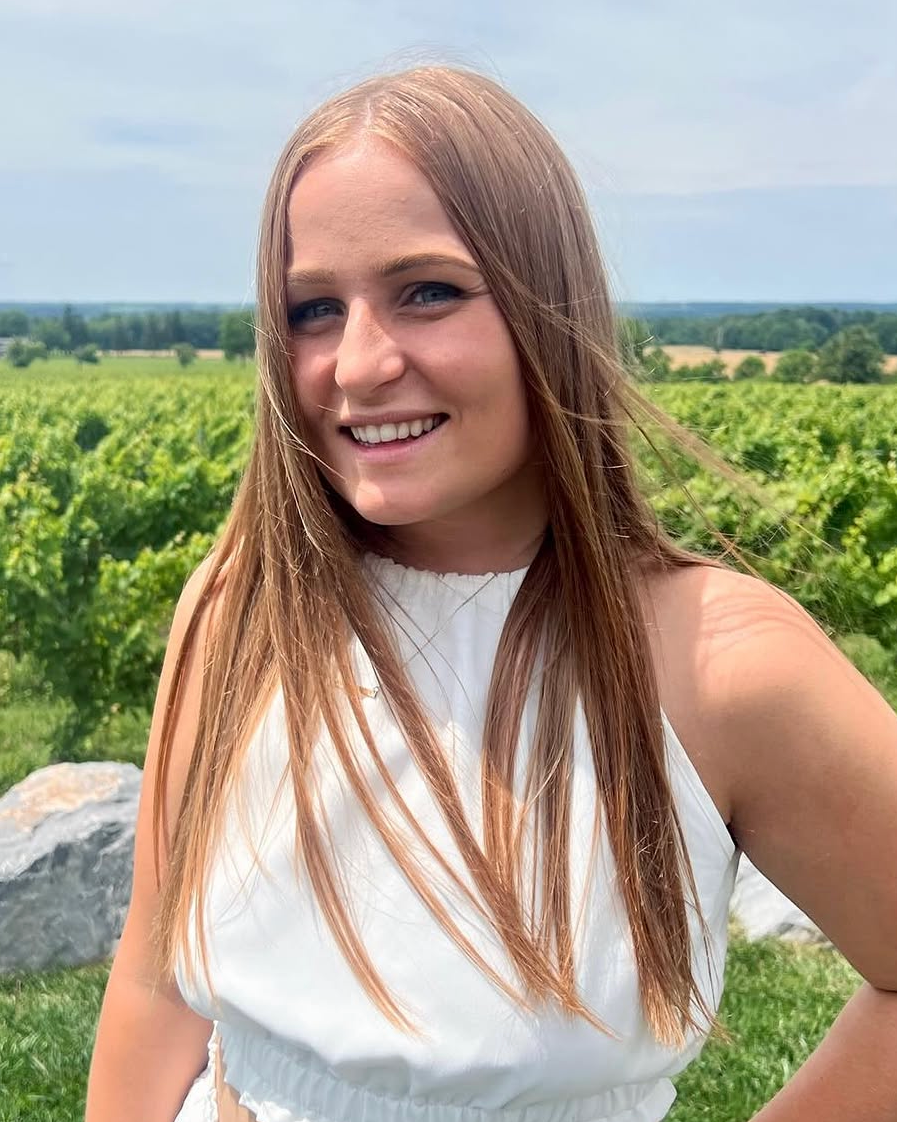 A young woman with long, straight, reddish-brown hair, smiling and taking a selfie outdoors in a rural area with green fields and trees in the background.