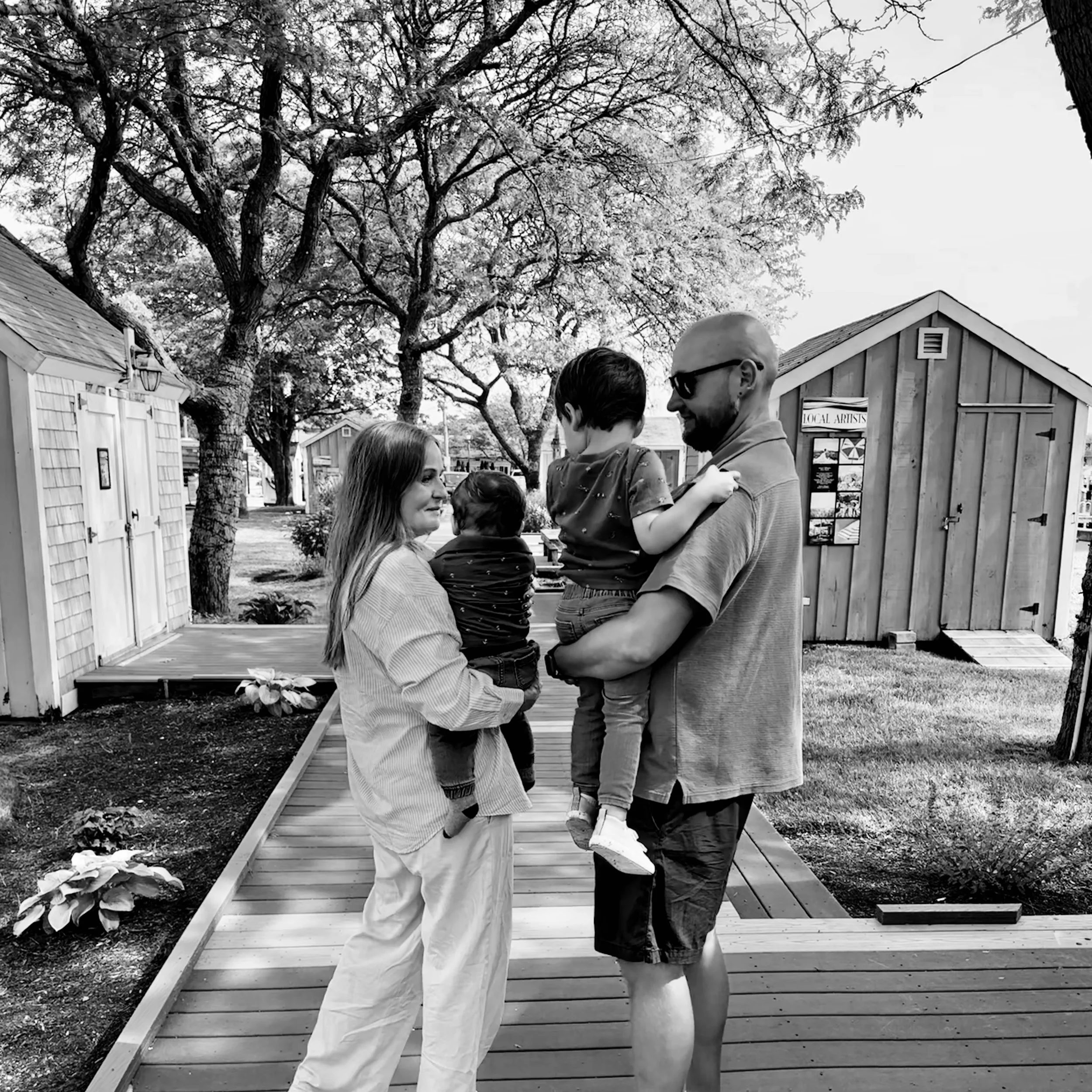 A black-and-white photo of two adults and two children standing on a wooden pathway outdoors, with large trees overhead and small buildings in the background.