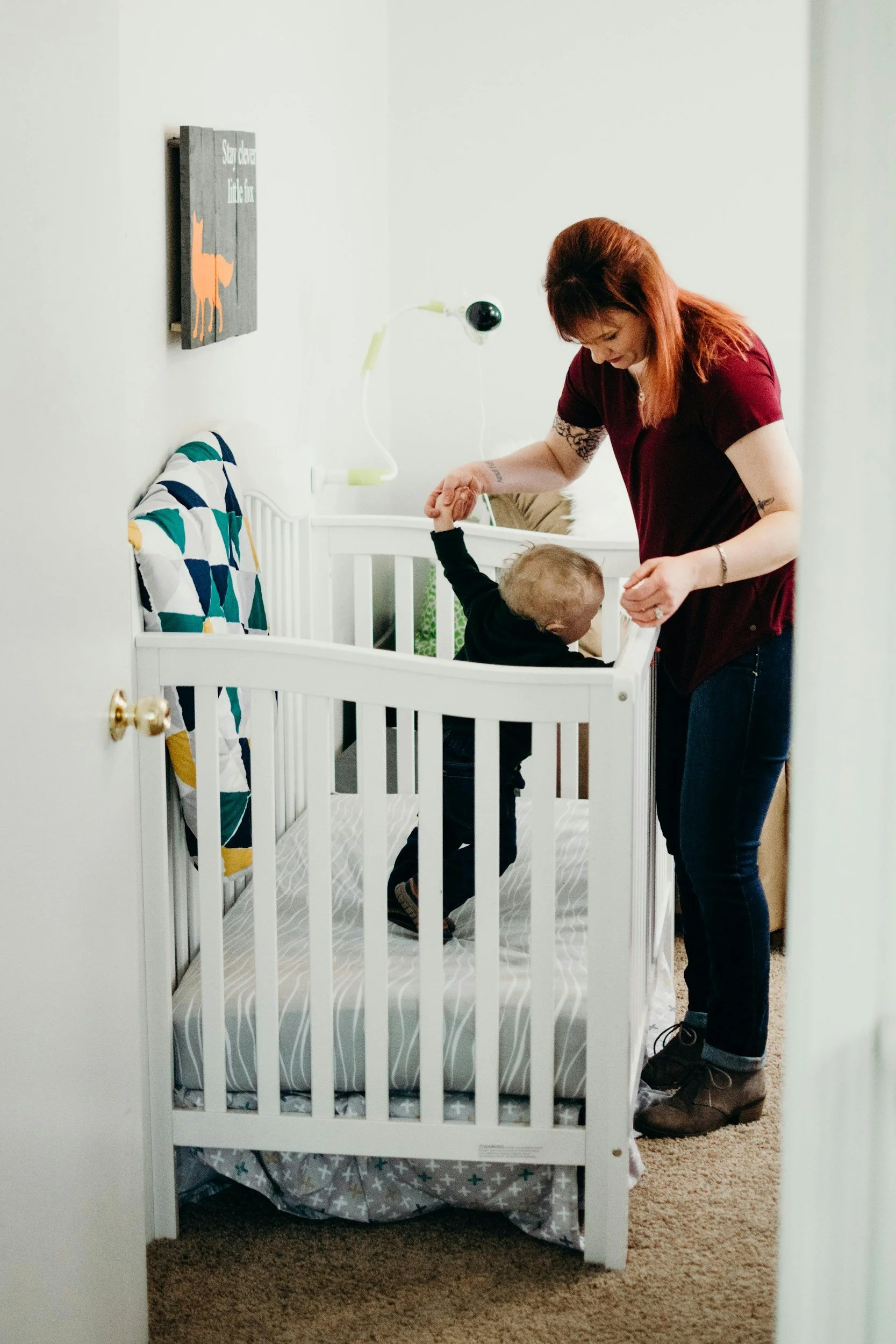 A woman helping a young child stand inside a white crib, with a bedside lamp and a wall-mounted sign that says 'Stay Calm, Little One'