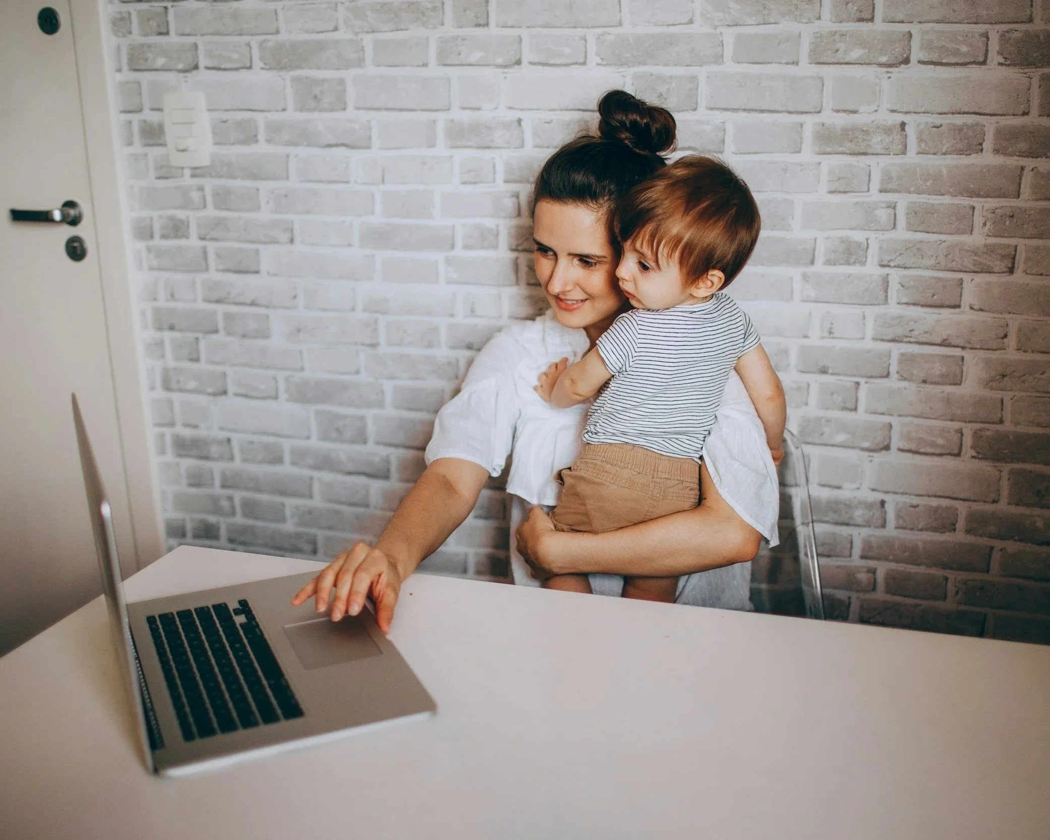 A woman holding a young boy while sitting at a table with a laptop in front of them, against a white brick wall background.