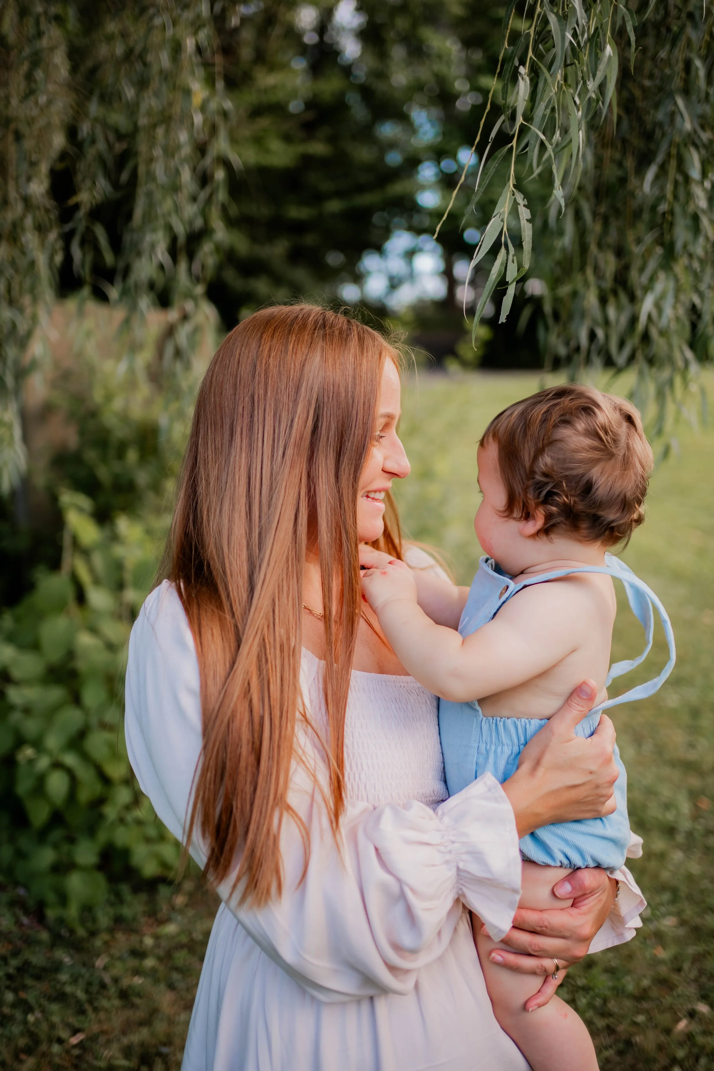 A woman with long red hair holding a young child with curly hair in an outdoor park setting. They are both smiling and looking at each other, with trees and greenery in the background.