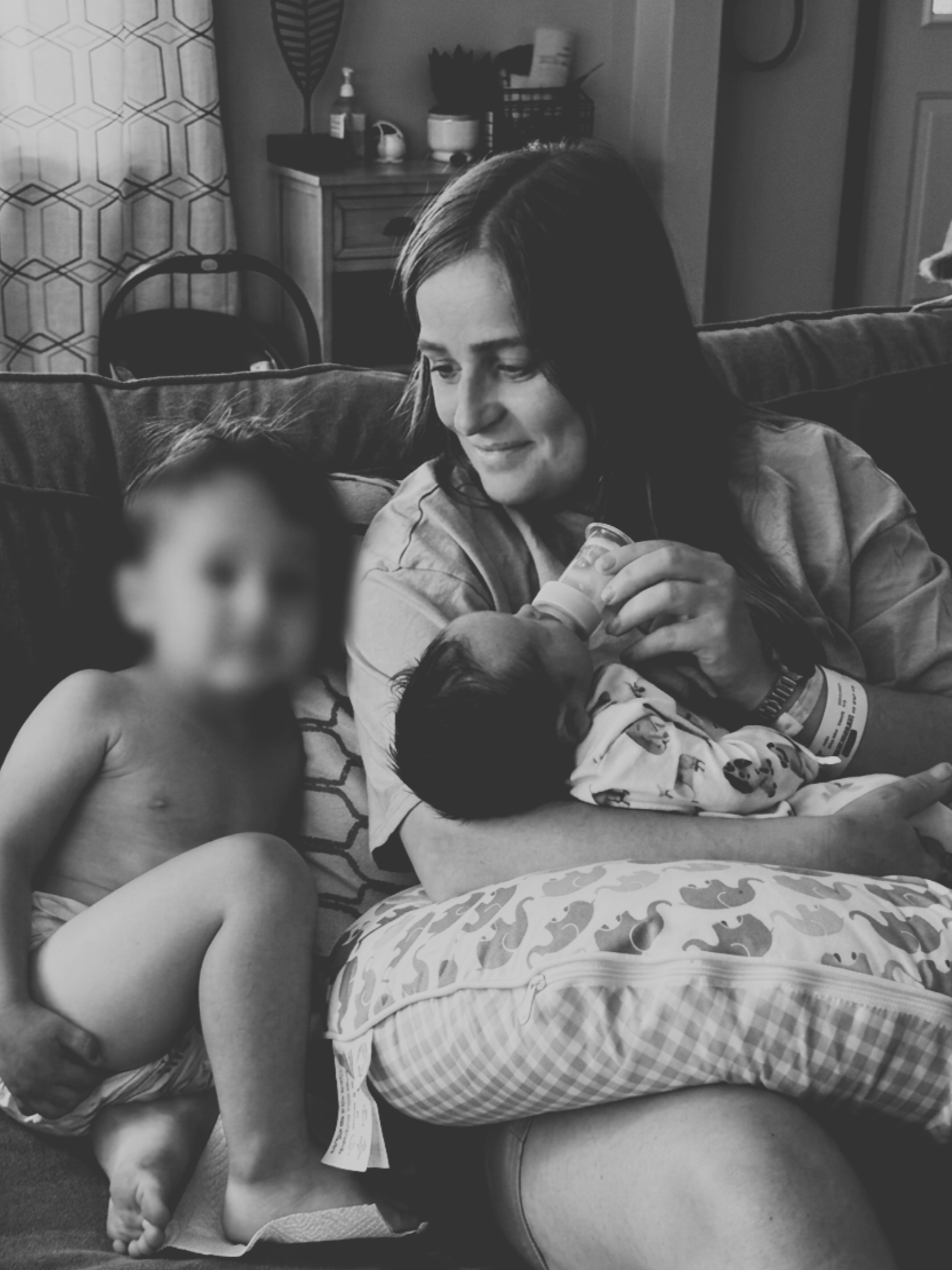 A woman sitting on a couch holding a baby, with a shirtless child sitting next to her. The woman is smiling and looking at the baby. In the background, there is a dresser with various items on top, including a plant, bottles, and a small appliance. The photo is in black and white.