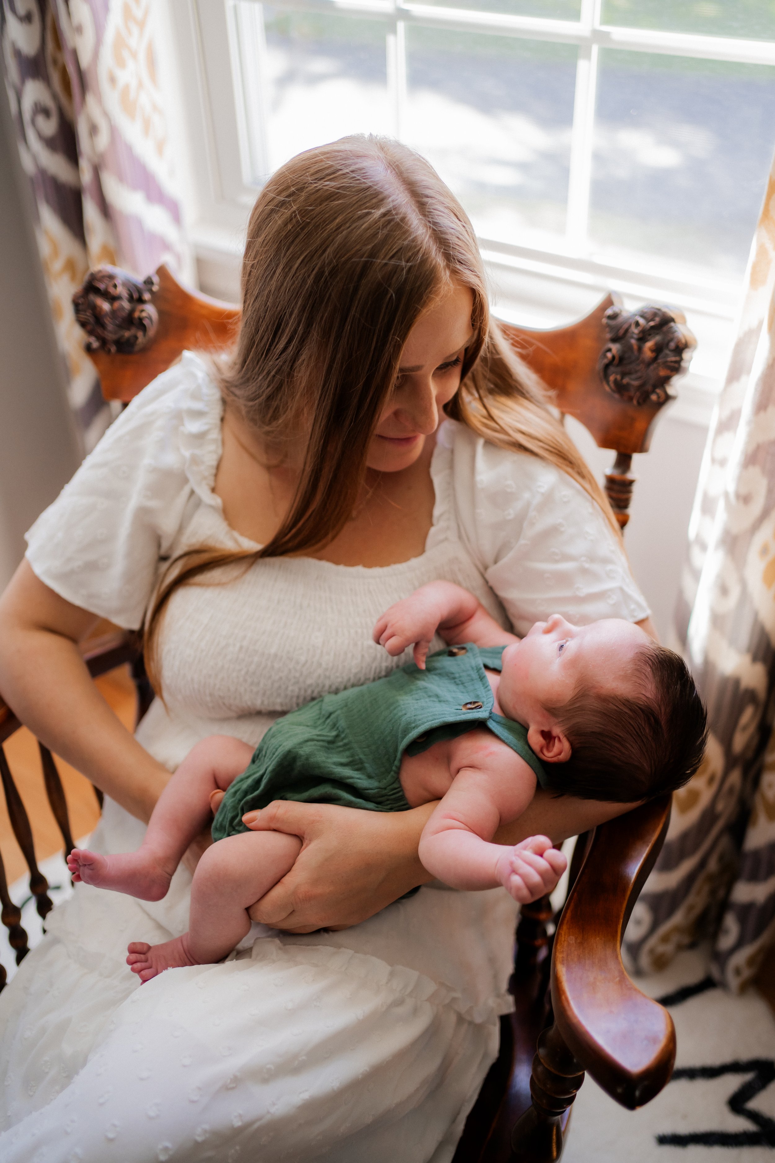 A woman with long reddish hair sitting on a wooden chair holding a sleeping baby with dark hair and light eyes, near a window with white curtains.