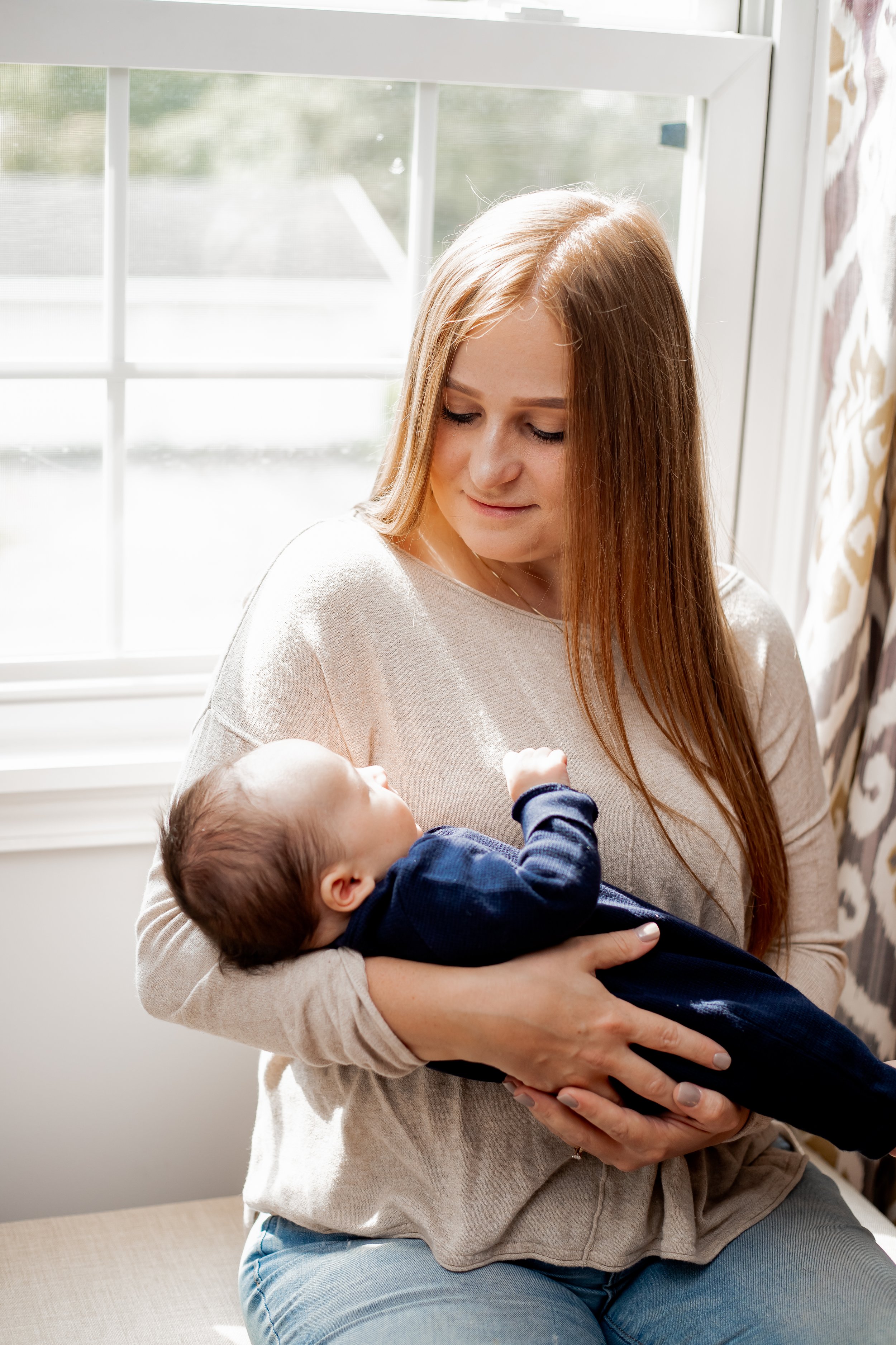 A woman with long red hair holding a baby with brown hair in a blue outfit, sitting by a window with white framing and light curtains, with natural sunlight illuminating the scene.